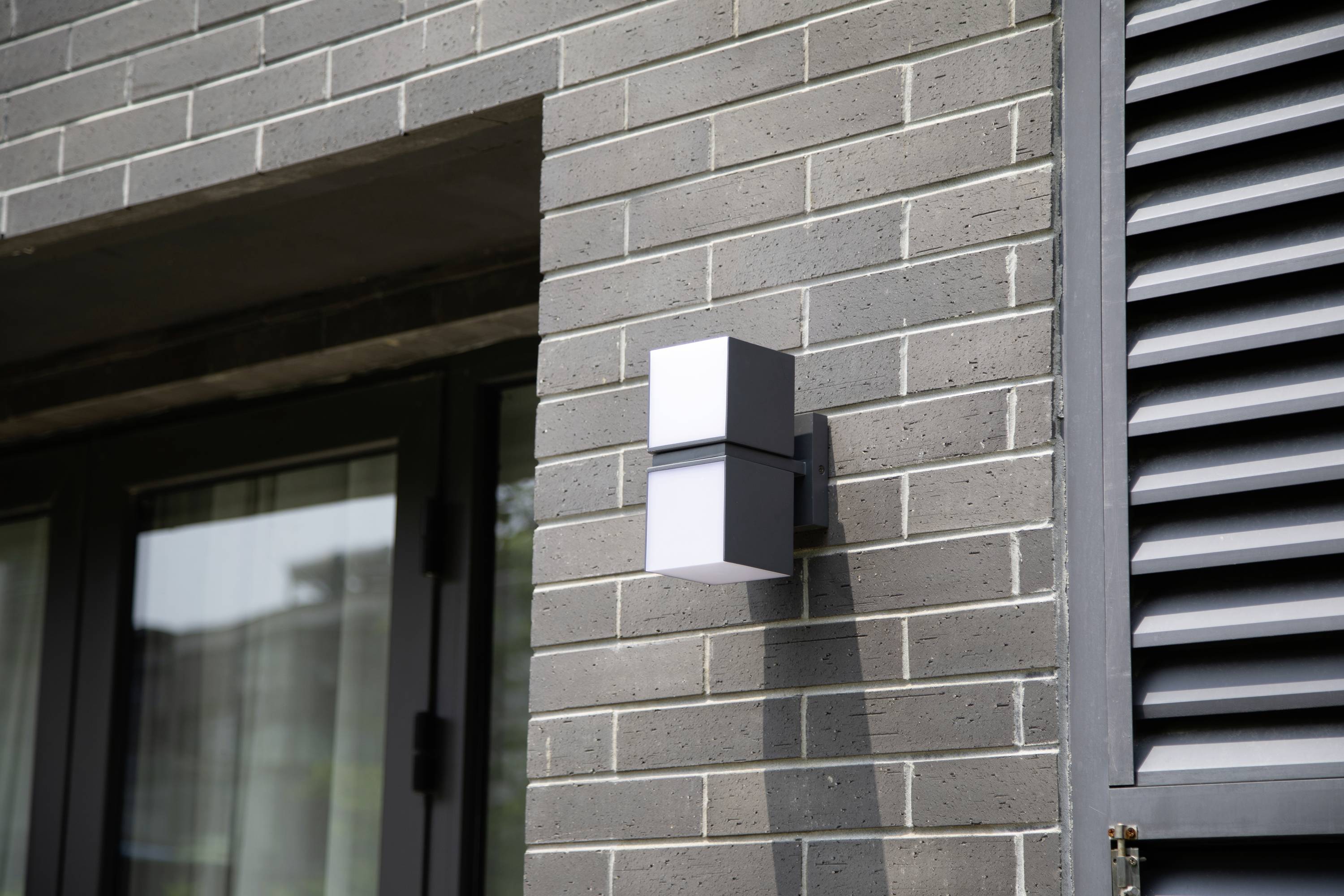 Un luminaire mural moderne est installé sur un mur de briques gris à côté d'une fenêtre et diffuse de la lumière dans les environs.