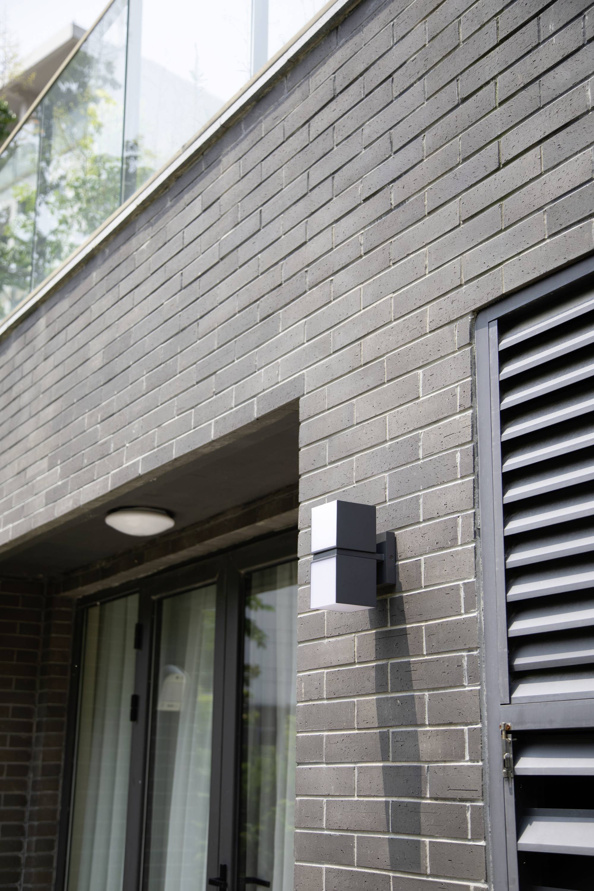 Façade moderne de maison avec des briques grises, une porte vitrée et un lampadaire extérieur noir. Un balcon en verre est visible en haut à gauche.