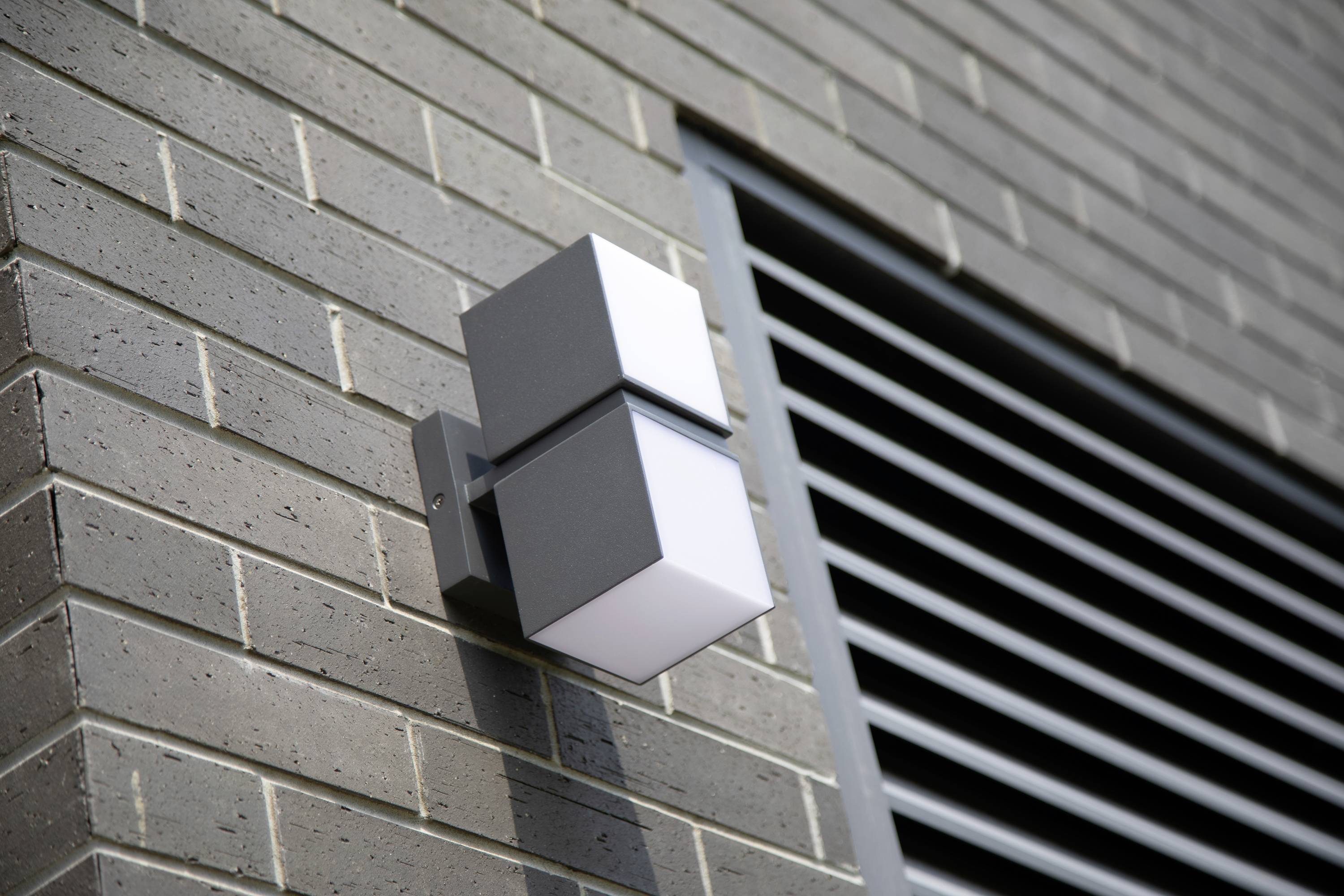 Applique murale extérieure de design moderne et cubique, montée sur un mur revêtu de briques. Des fentes d'aération sont visibles sur le mur.