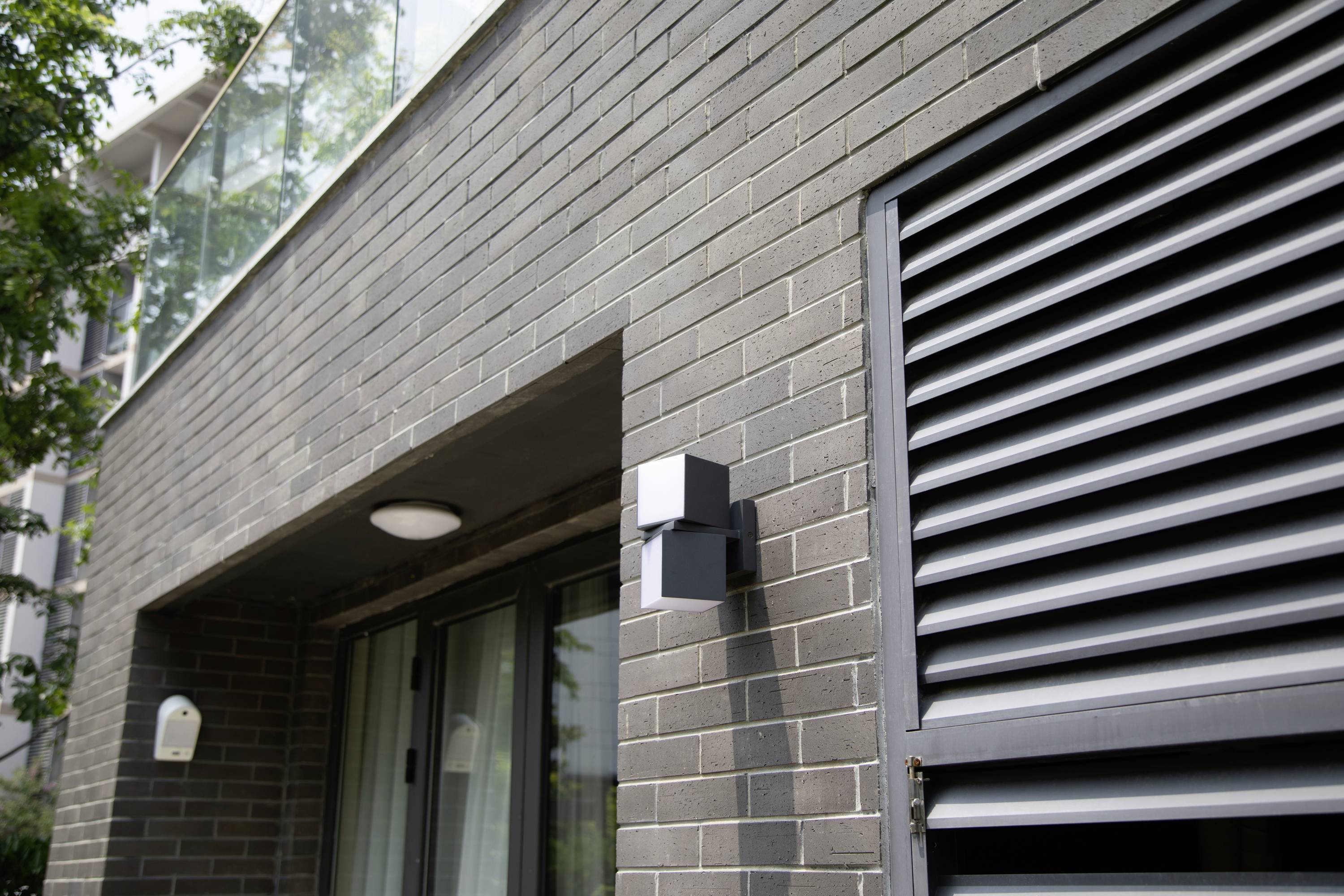 Vue extérieure d'un bâtiment moderne avec des briques grises, une grande baie vitrée, un éclairage mural noir et des grilles de ventilation.