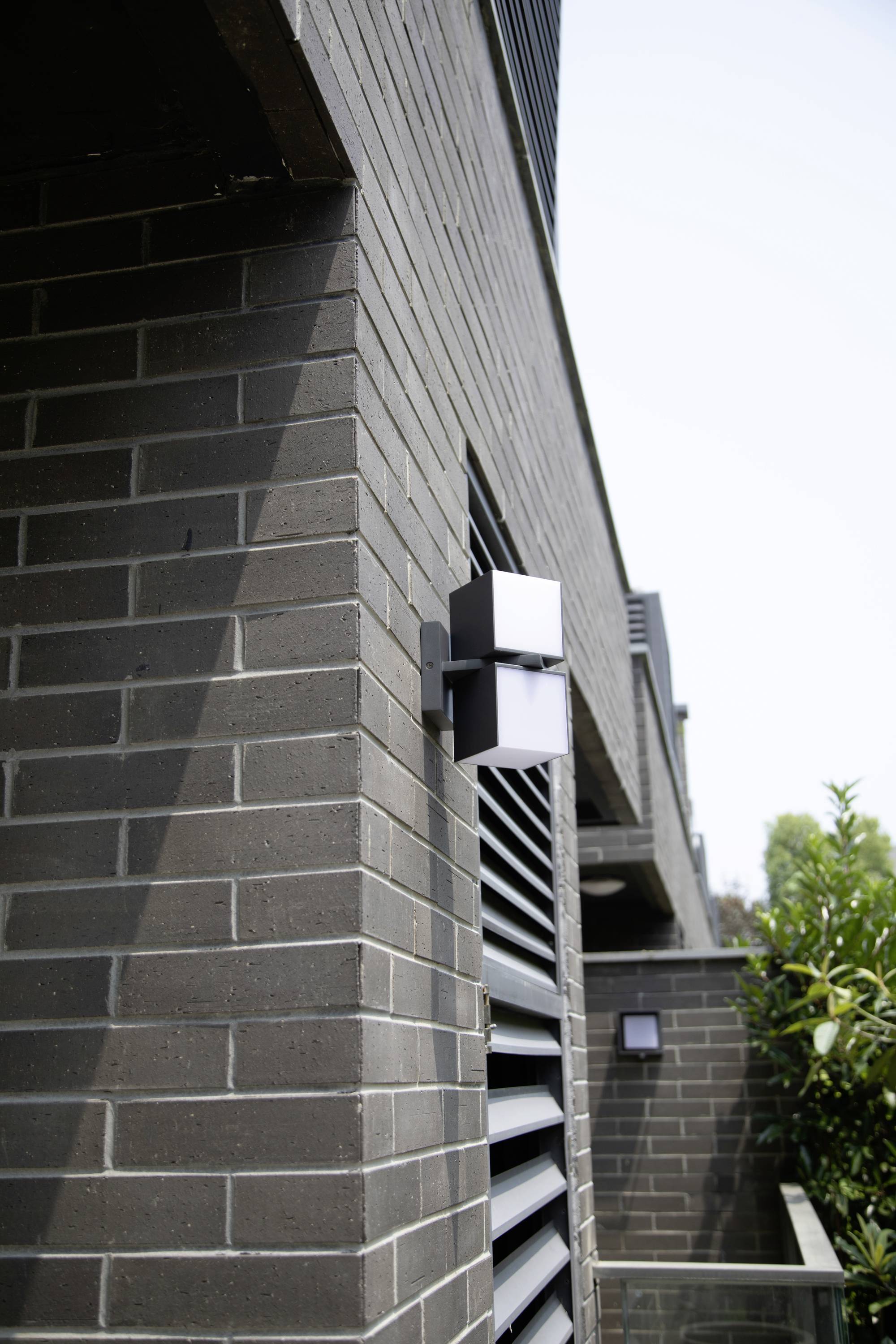 Bâtiment moderne avec façade en briques de couleur gris foncé et luminaire extérieur géométrique. Une autre lumière est visible en arrière-plan.