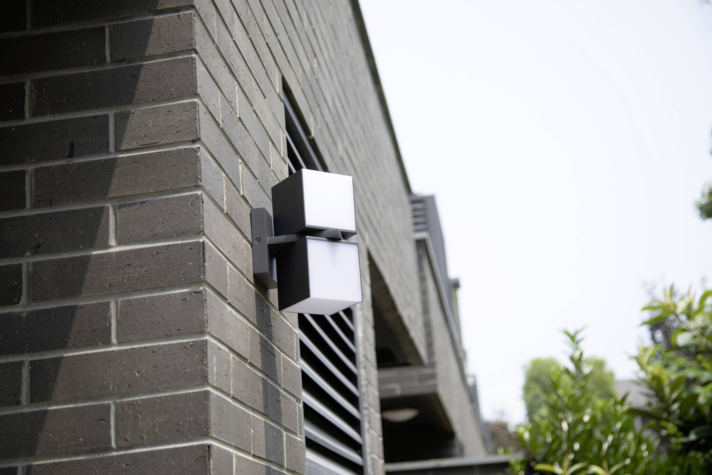 Lampe extérieure moderne fixée sur un mur de briques d'un bâtiment, entourée de plantes vertes en arrière-plan.