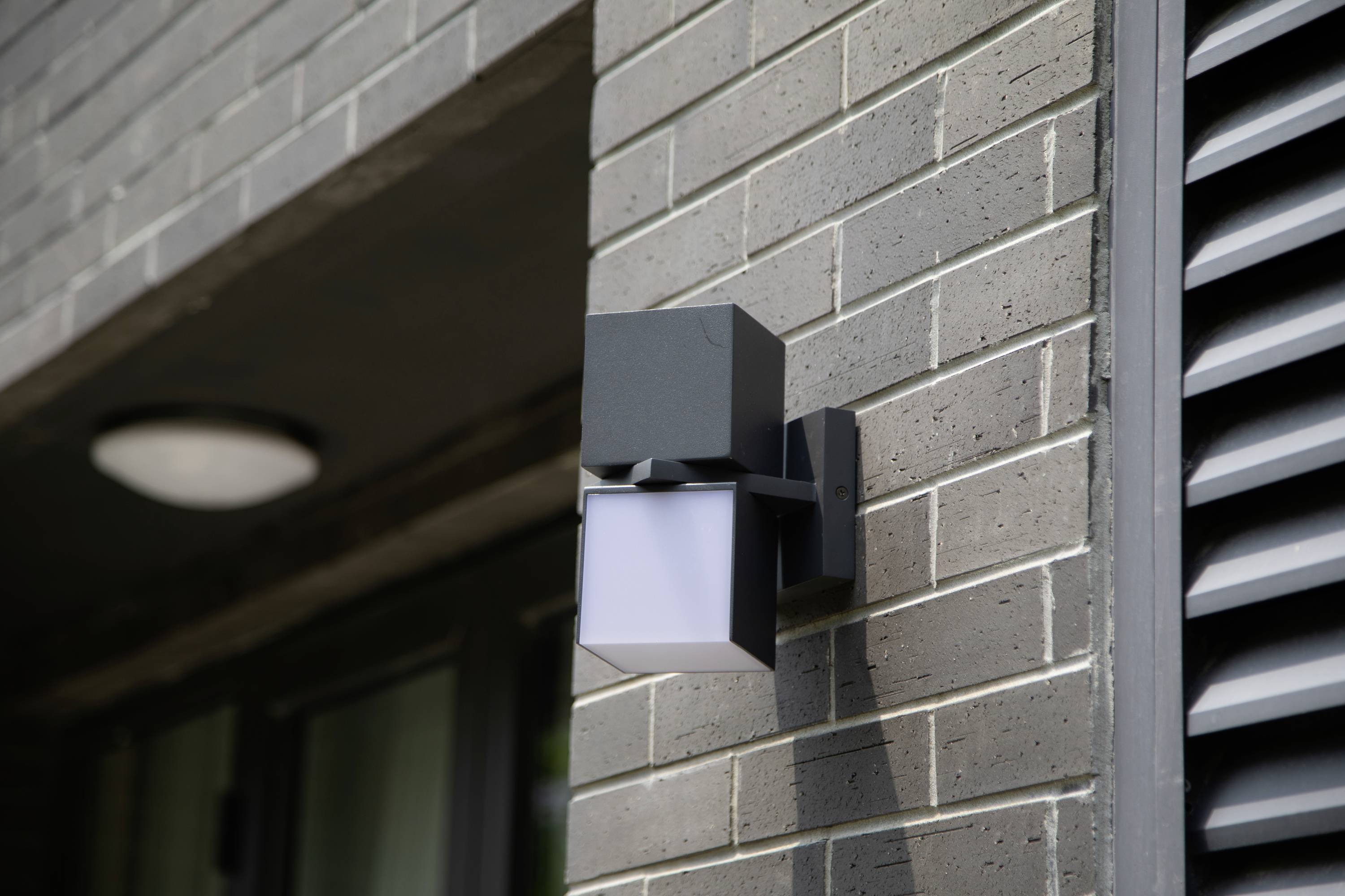 Applique extérieure montée sur un mur de briques grises. La lampe présente un design moderne et carré, illuminant la zone d'entrée.