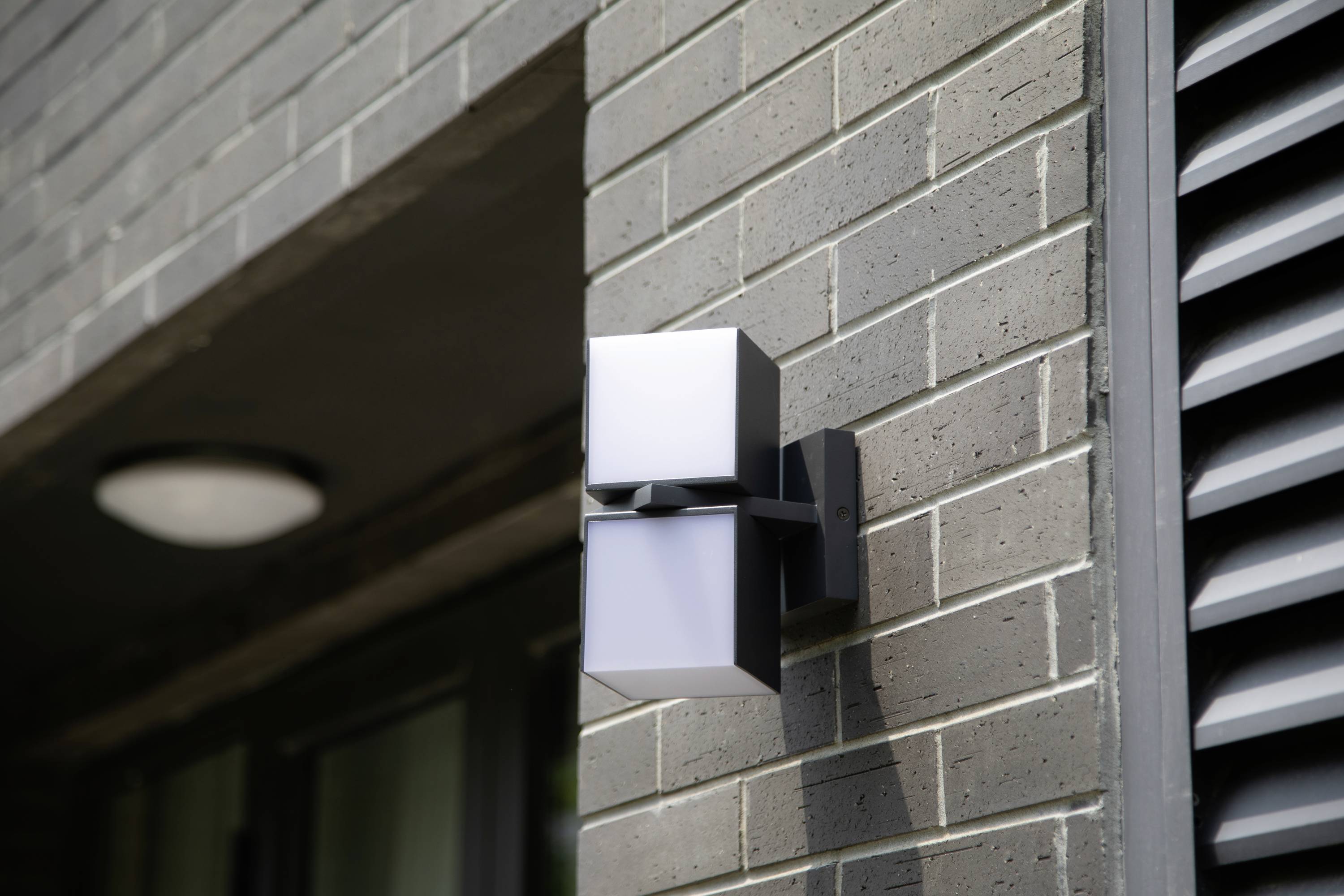 Un luminaire extérieur moderne est installé sur un mur en briques grises. Il présente un design minimaliste et éclaire dans deux directions.