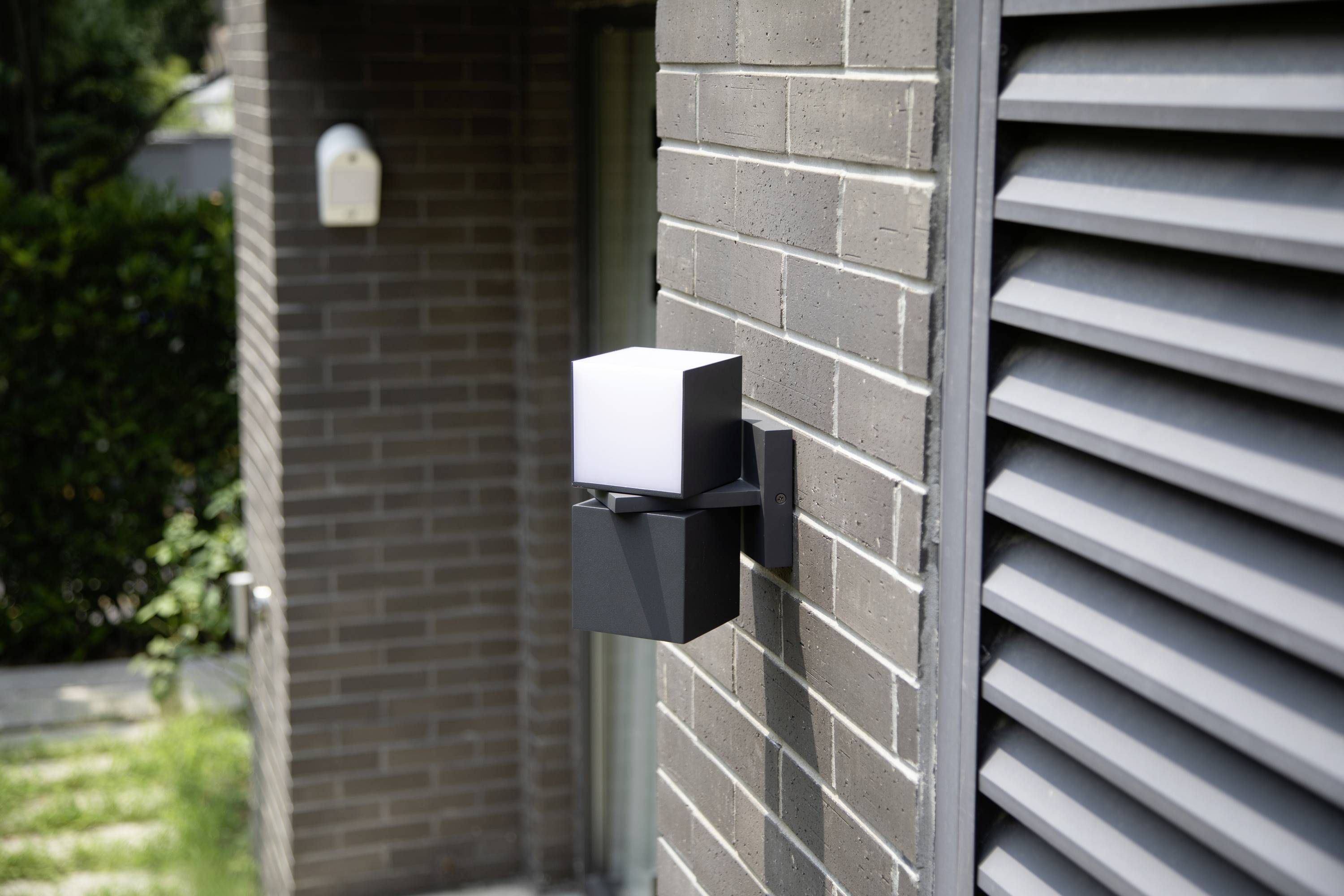 Applique murale au design moderne et cubique sur un mur de briques. Un jardin avec un espace vert est visible en arrière-plan.