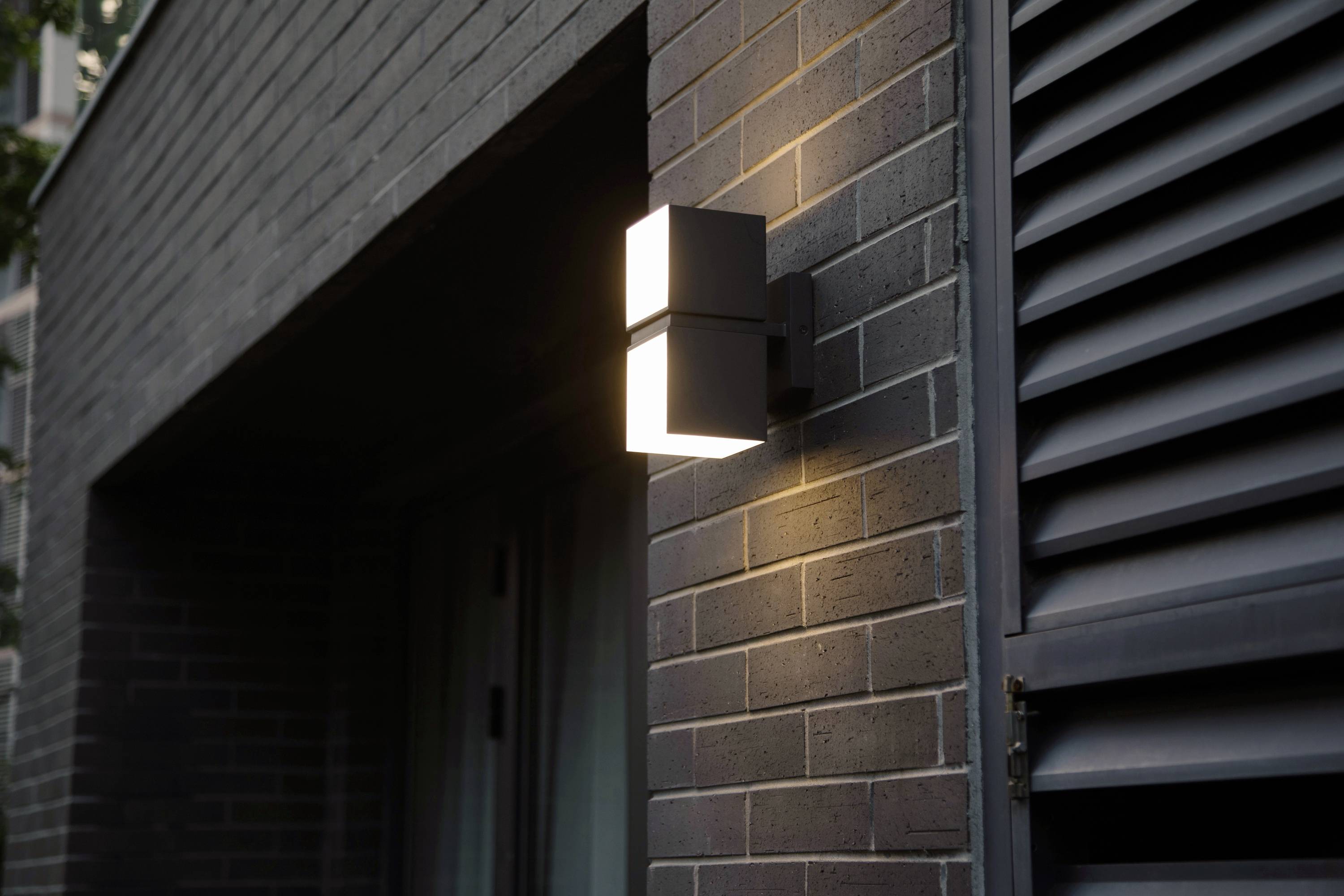 Une lampe extérieure moderne éclaire le mur de la maison au crépuscule, offrant un éclairage et une sécurité pour l'entrée.