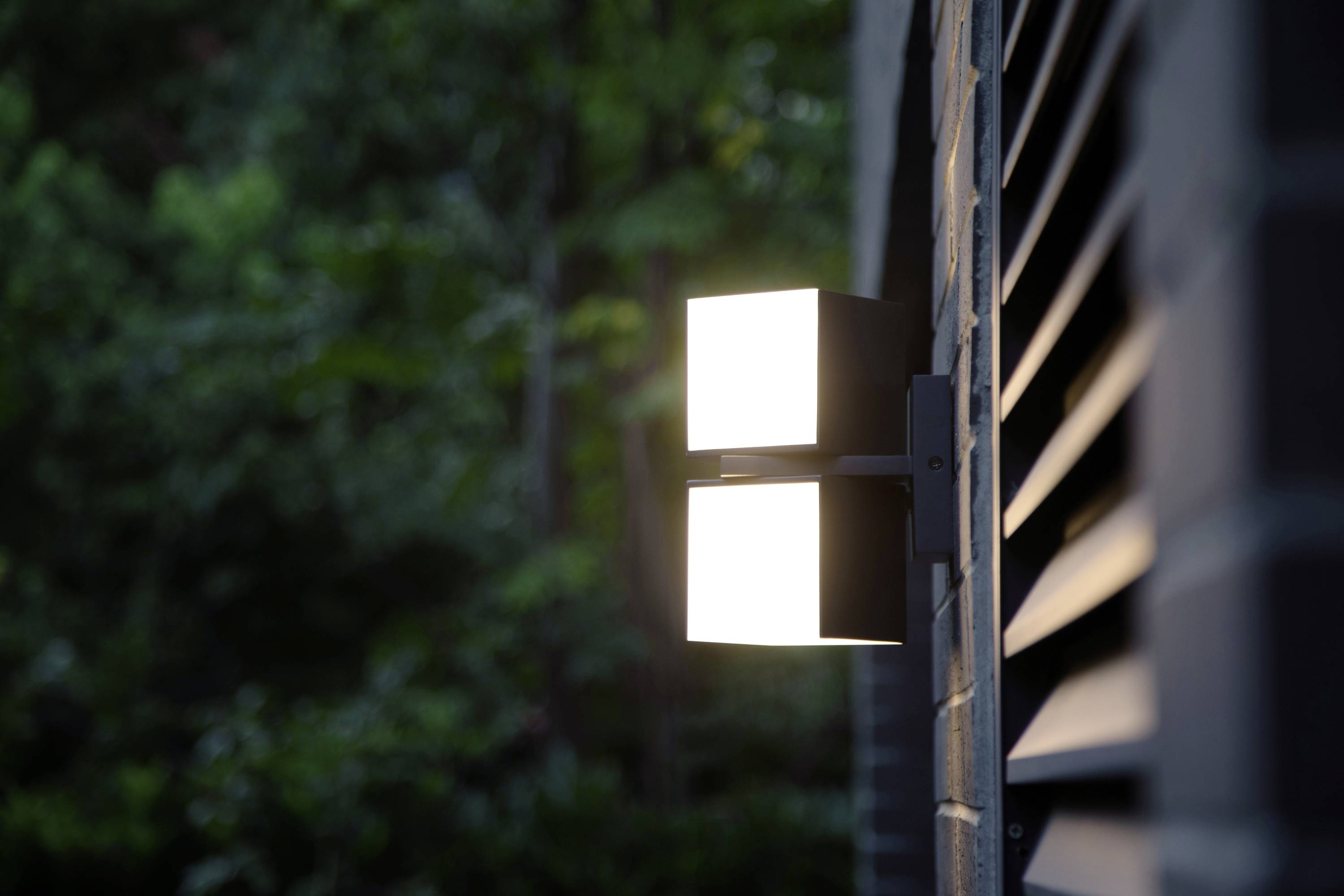 Lampe extérieure moderne fixée à un mur de briques éclairant une zone de jardin. Des feuilles vertes floues sont visibles en arrière-plan.