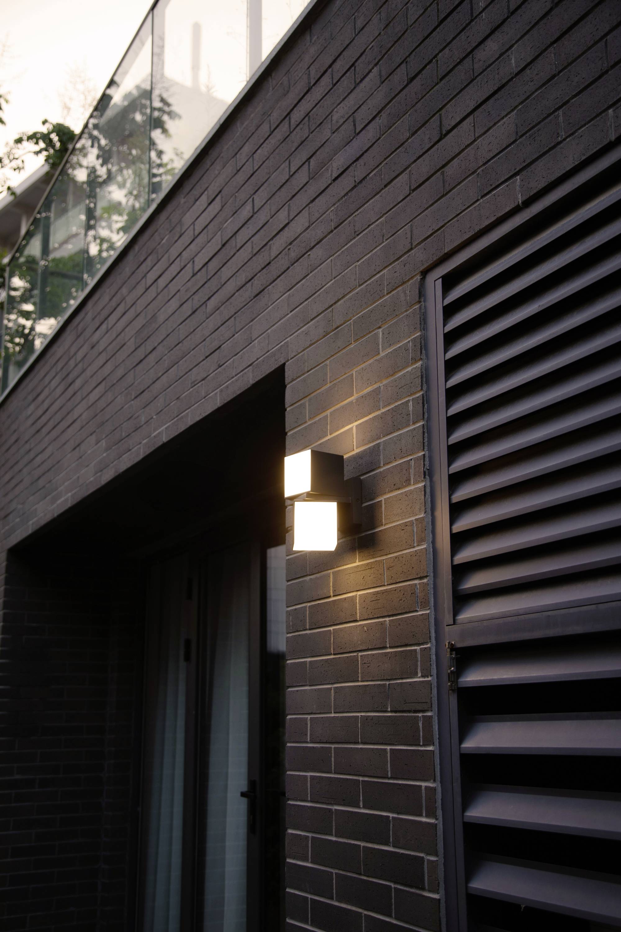 Un éclairage extérieur moderne sur un mur de briques à côté d'une porte, illuminant la zone au crépuscule.