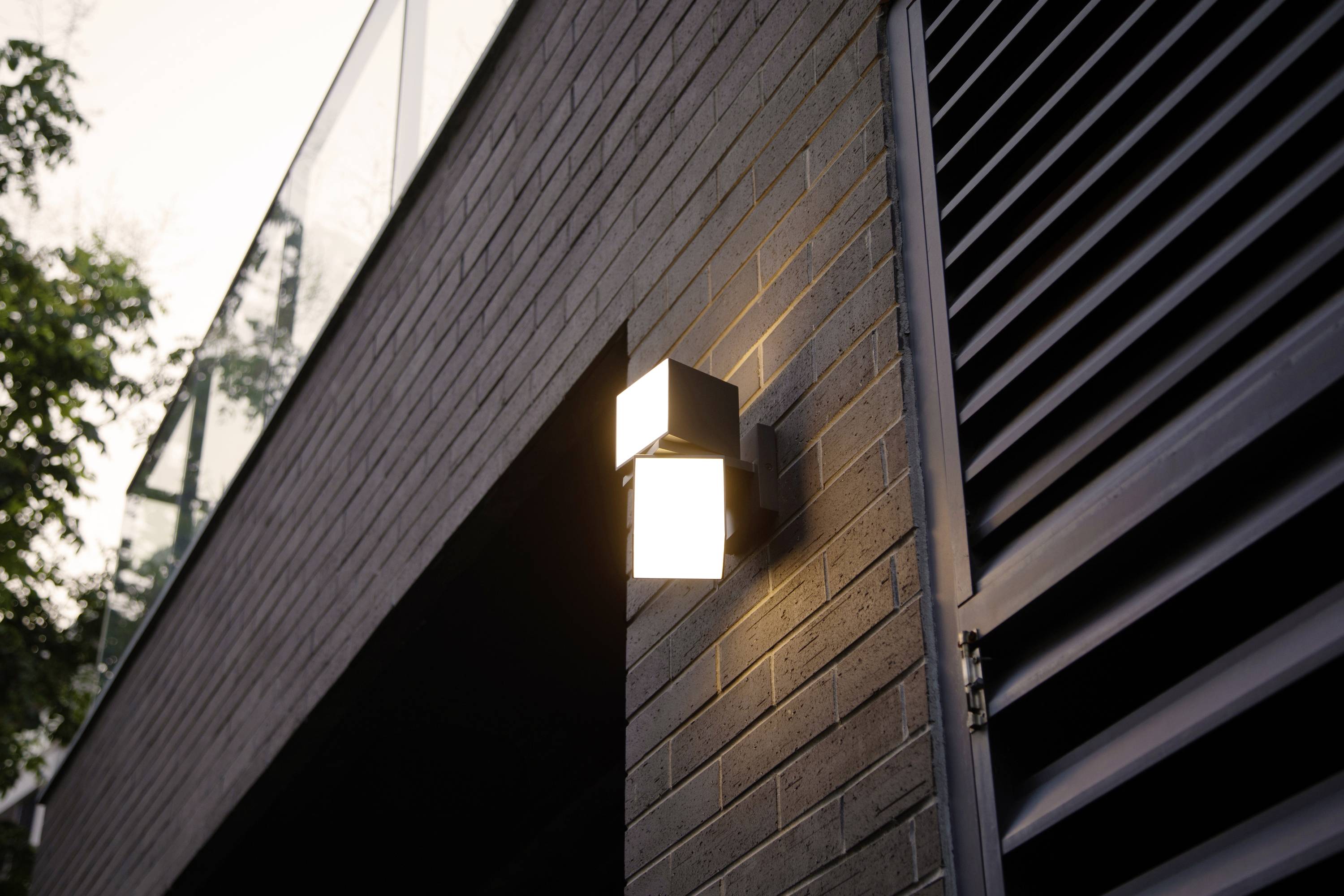 Applique murale éclairée sur une façade de briques au crépuscule. À l'arrière-plan, on aperçoit une balustrade en verre et un arbre.
