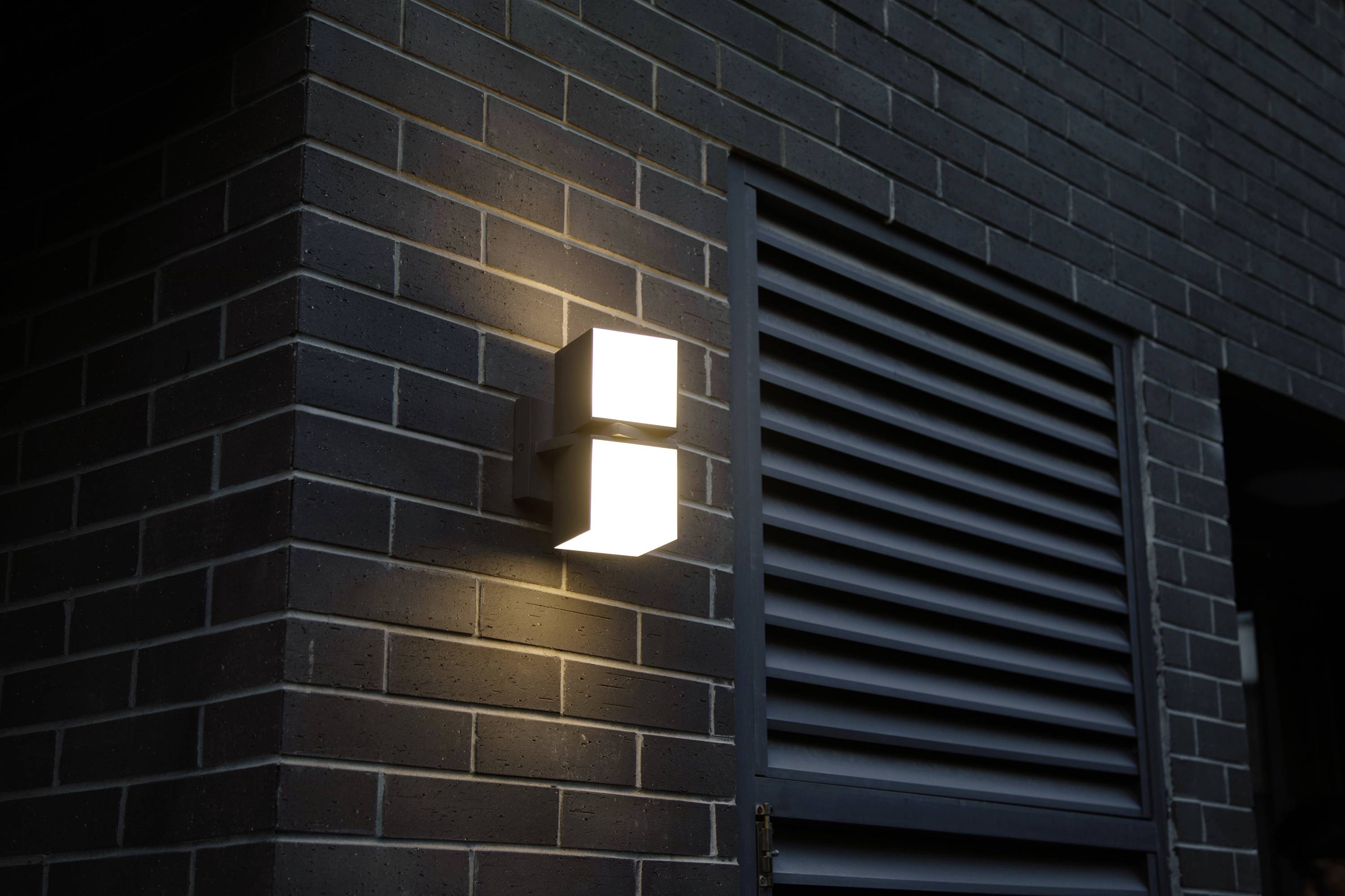 Un luminaire moderne projette une lumière chaleureuse sur un mur revêtu de briques sombres, à côté d'une fenêtre équipée de stores.