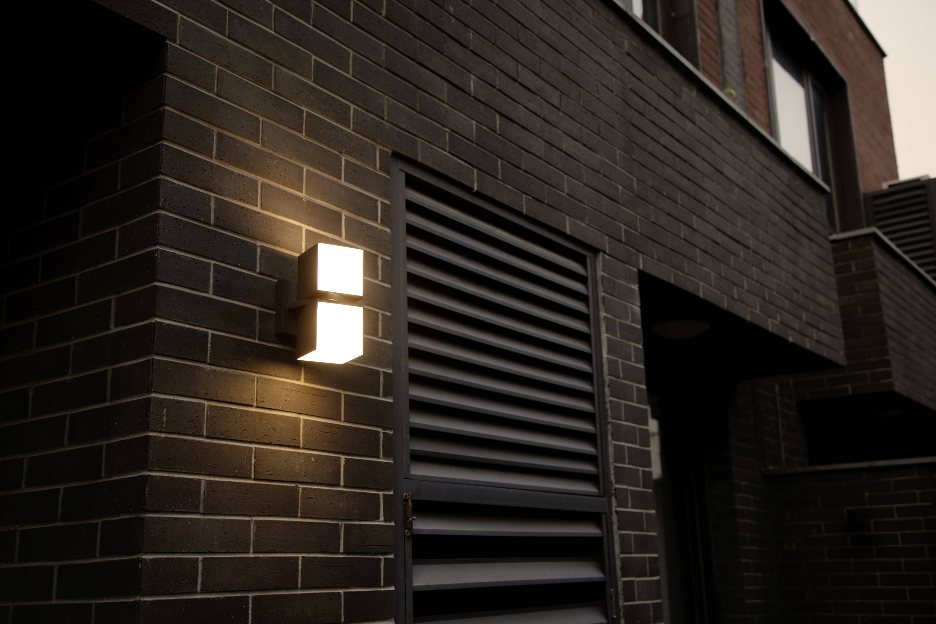 Façade d'un bâtiment moderne en briques sombres avec un dispositif d'éclairage mural allumé, diffusant une lumière douce.