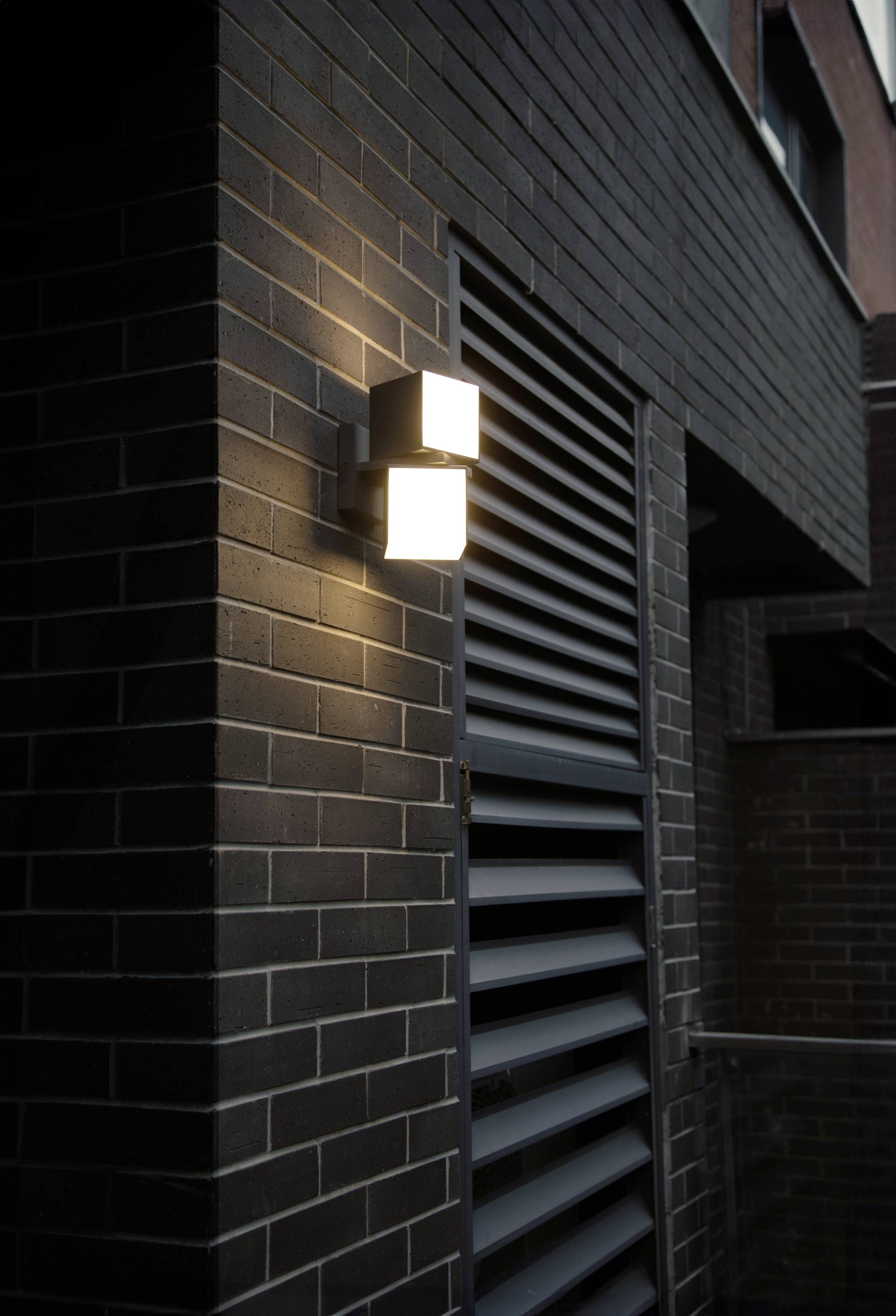 Luminaire extérieur sur une façade en briques sombres, allumé la nuit et créant un effet de lumière chaleureux et subtil.