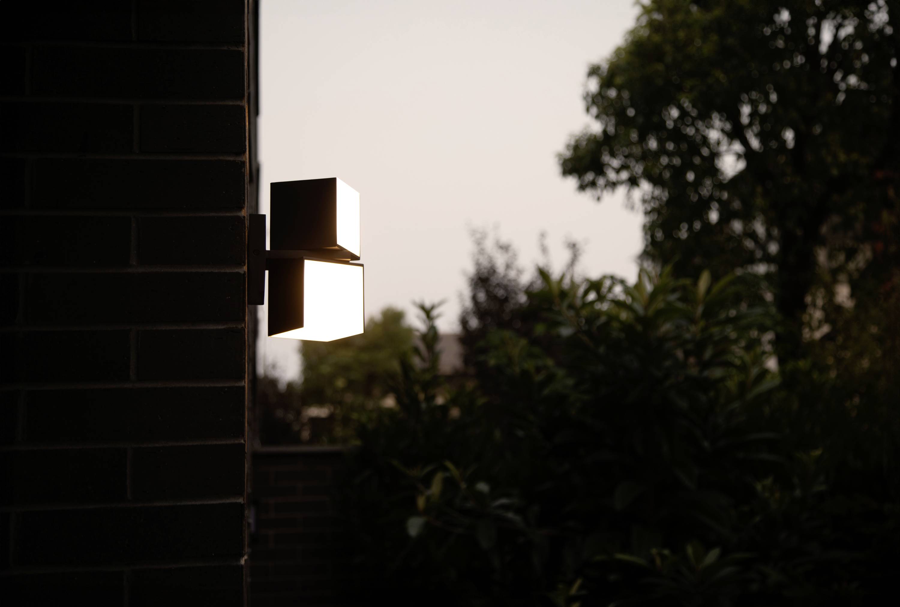 Éclairage extérieur sur un mur de maison au coucher du soleil, brillant dans le crépuscule, avec des arbres en arrière-plan.