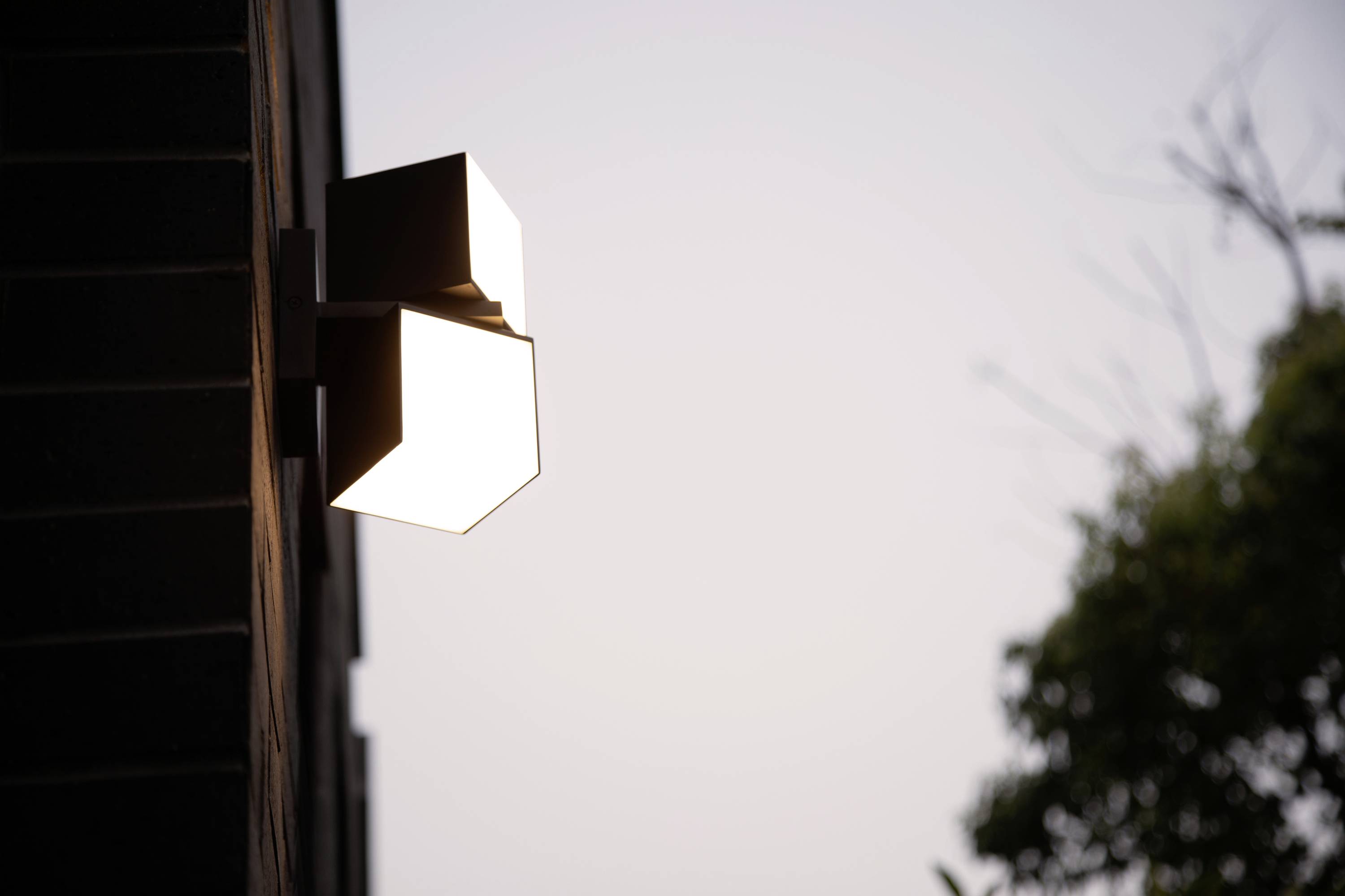 Une lampe extérieure moderne fixée au mur d'une maison éclaire au crépuscule. En arrière-plan, on aperçoit des arbres et un ciel dégagé.