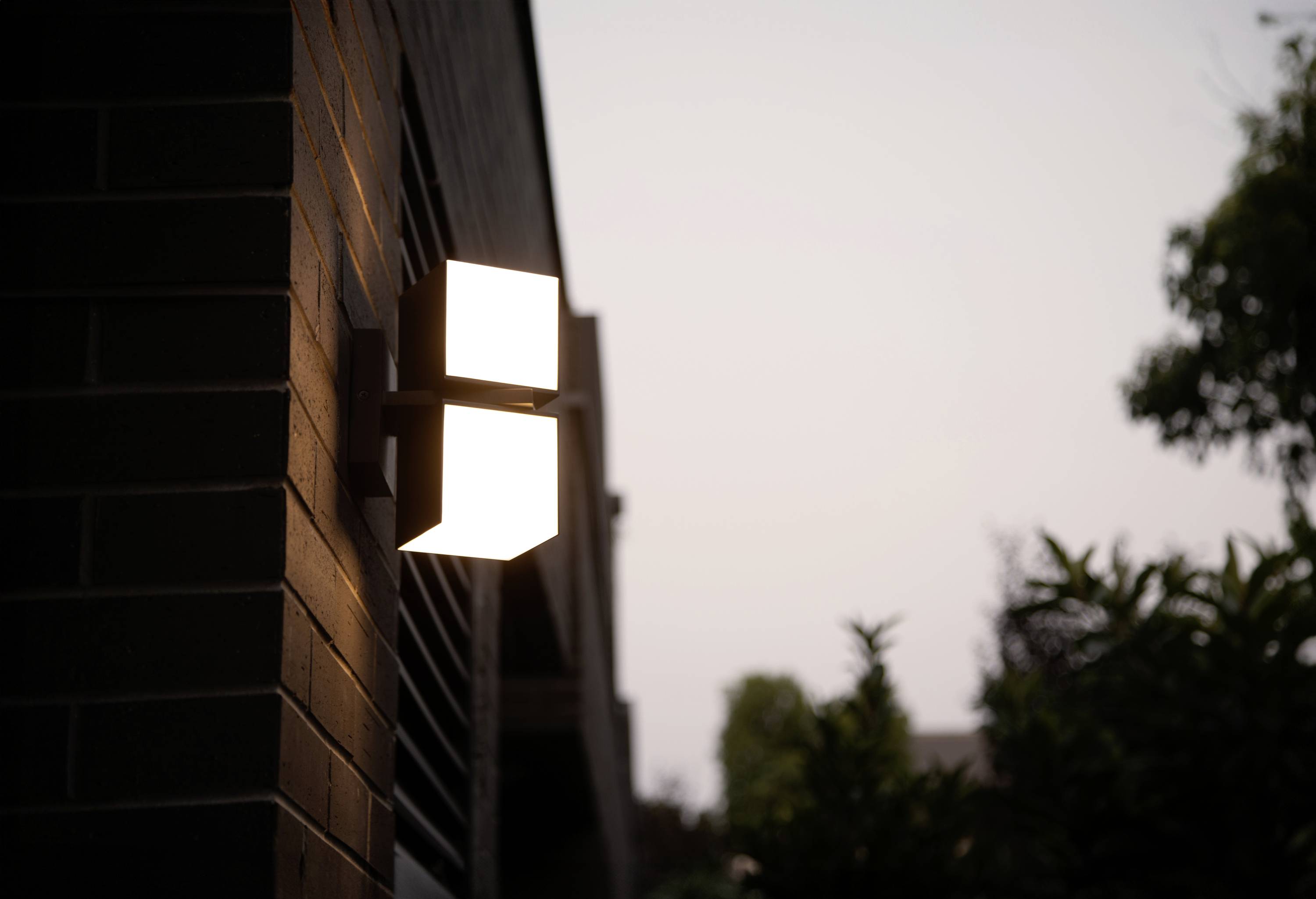 Éclairage extérieur sur un mur de maison au crépuscule. La lampe diffuse sa lumière vers le haut et vers le bas, illuminant doucement la brique.