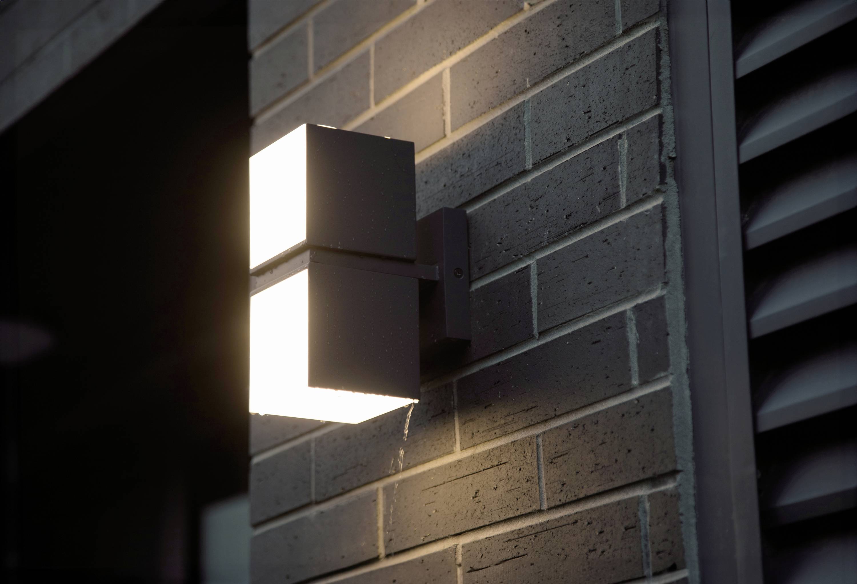 Une lampe extérieure moderne éclaire un mur de briques et diffuse une lumière chaleureuse au crépuscule.