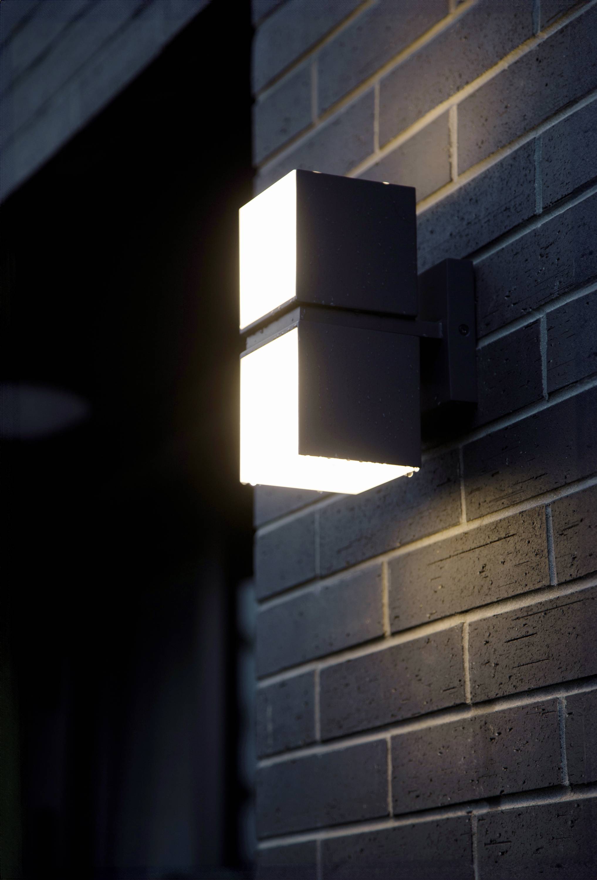 Applique murale extérieure au design moderne et cubique, fixée sur un mur de briques, illuminant l'environnement au crépuscule.
