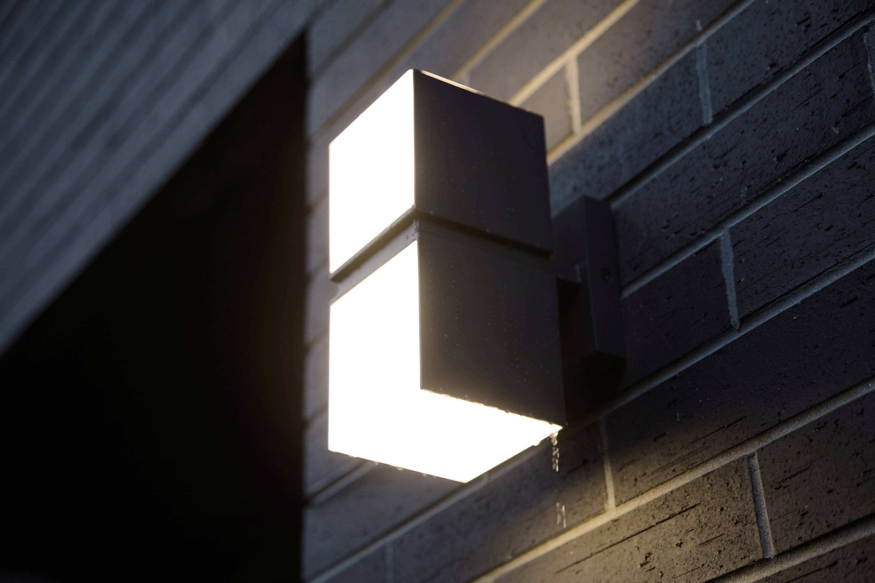 Lampe murale extérieure au design rectangulaire et moderne, illuminant un mur de briques gris foncé.