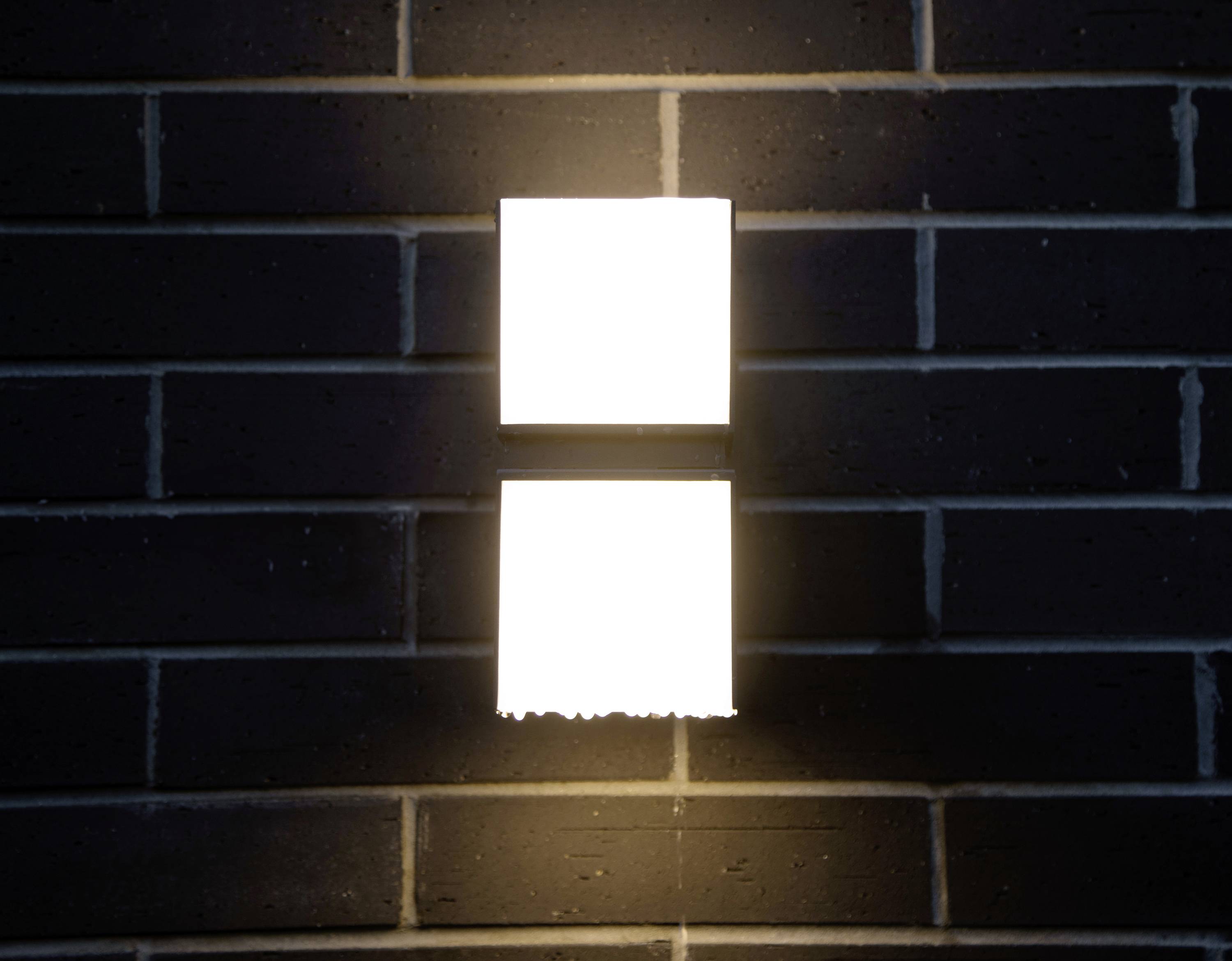 Deux luminaires carrés lumineux disposés verticalement sur un mur de briques sombres.