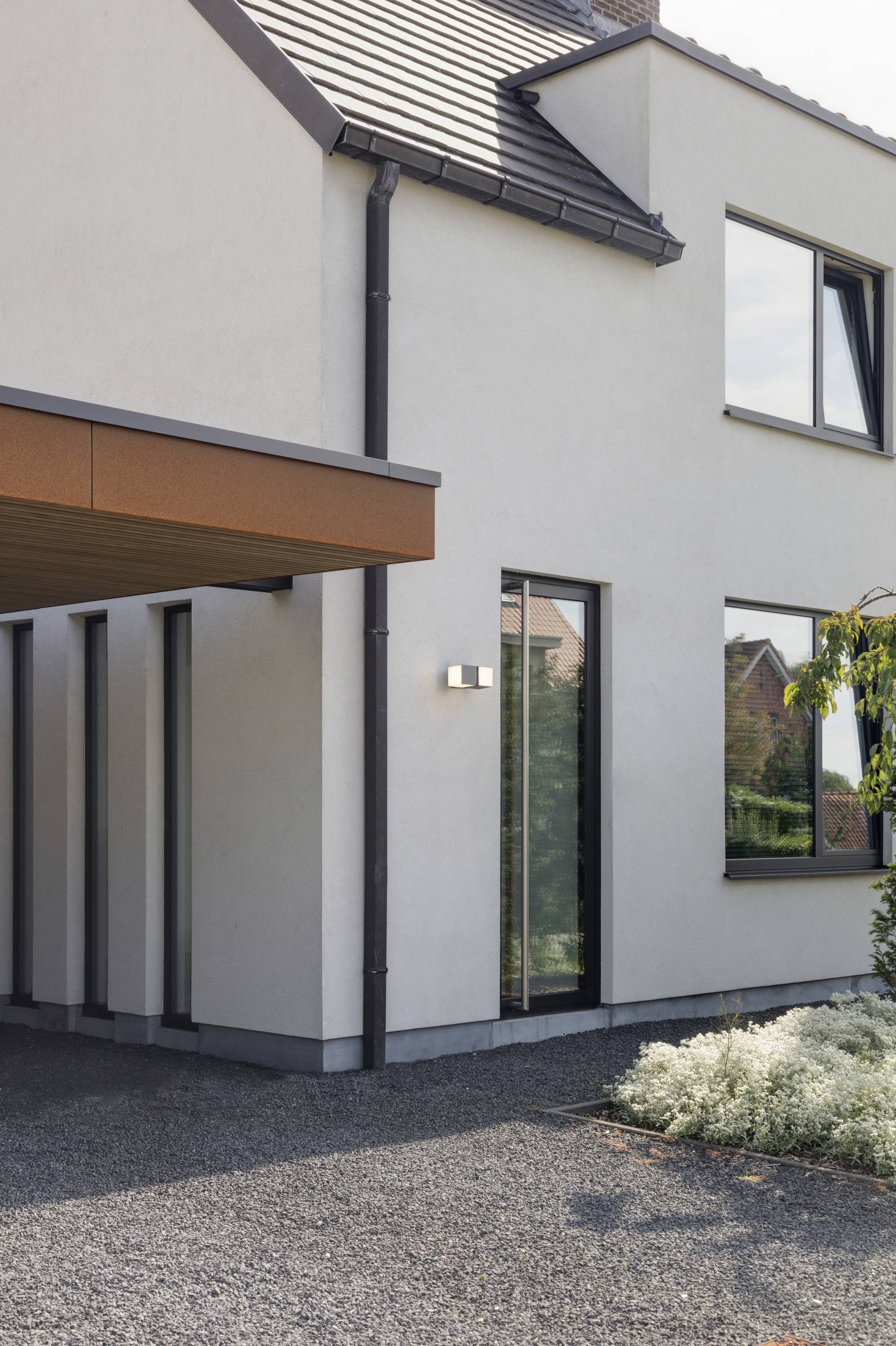 Maison moderne à deux étages avec des murs blancs et de grandes fenêtres. Un carport est construit sur le côté. Des arbustes sont plantés devant la maison.