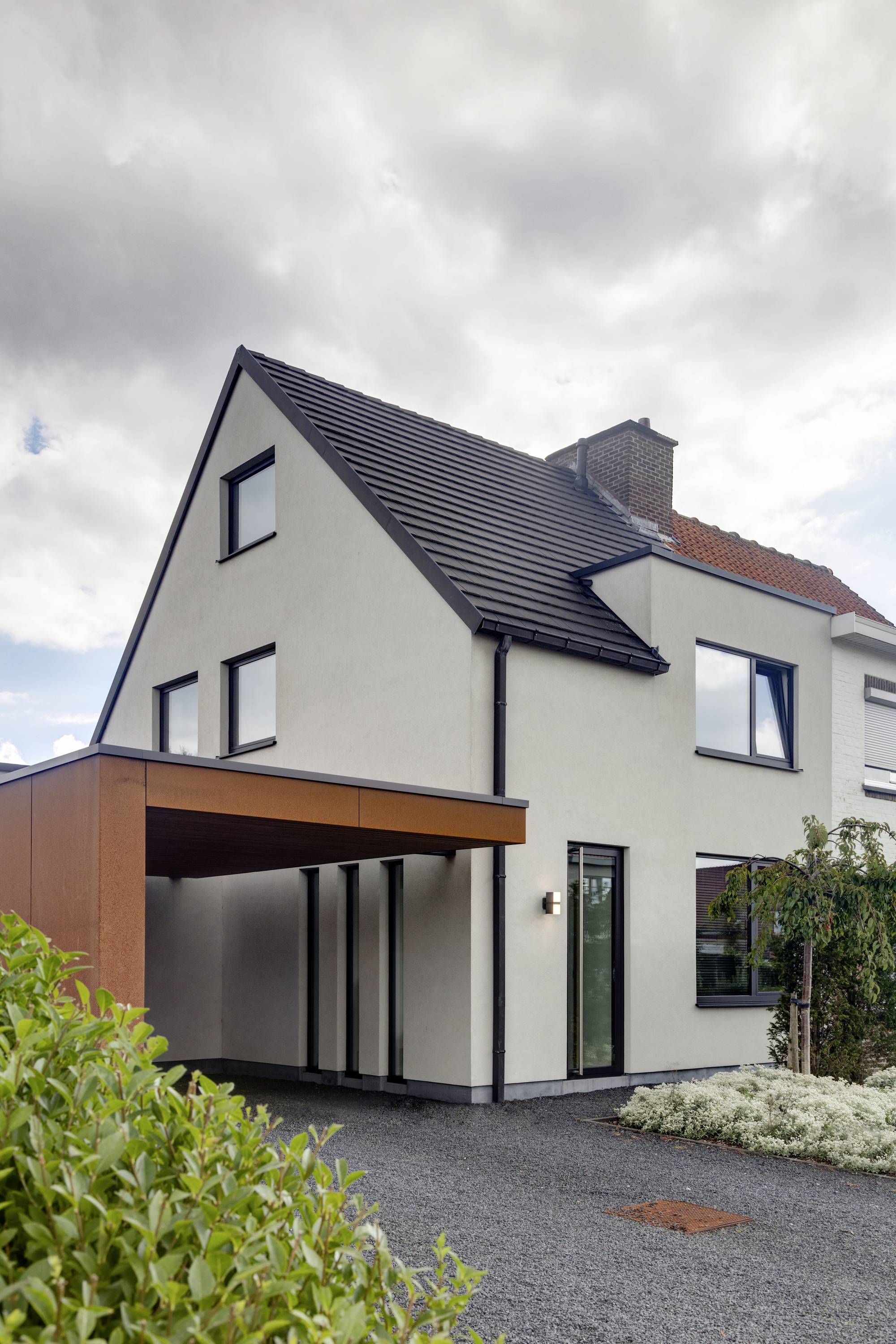 Une maison individuelle moderne avec une façade blanche, des encadrements de fenêtres noirs et un carport en bois. Devant la maison se trouvent des arbustes et un chemin de gravier.