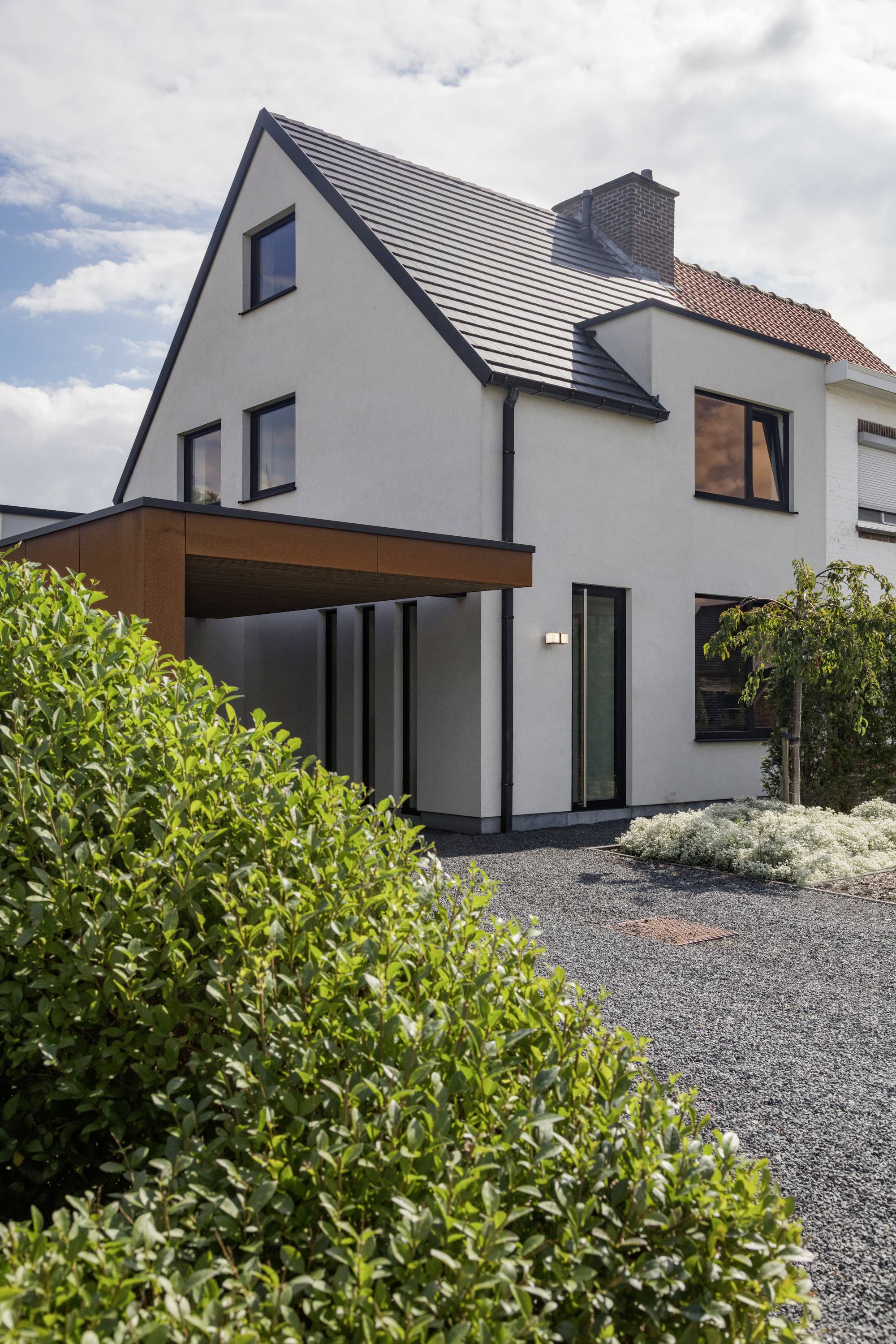 Maison moderne avec toiture à deux pans et façade blanche, entourée de buissons verts et d'un chemin de gravier, sous un ciel nuageux.
