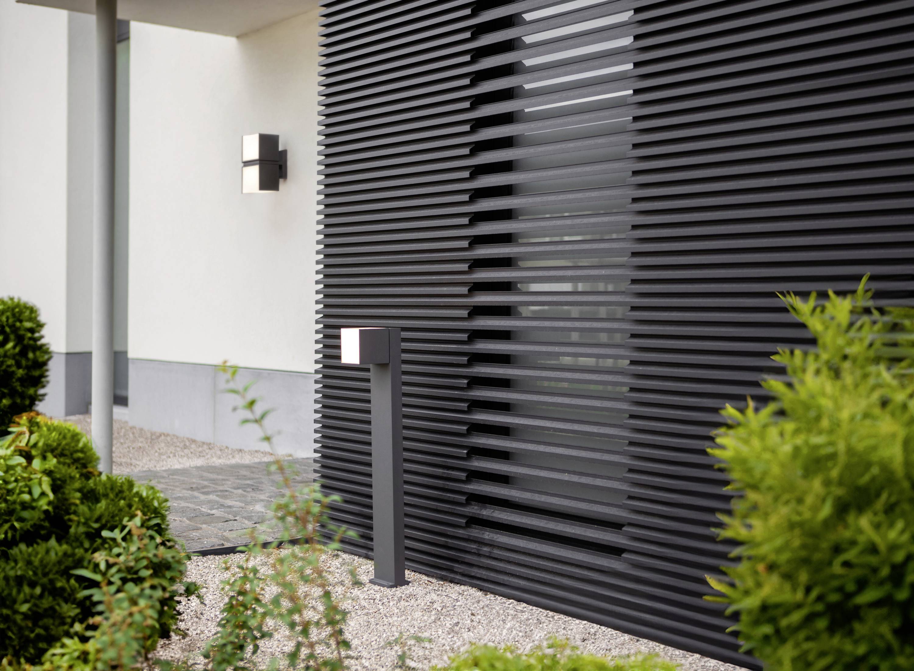 Façade moderne d'un bâtiment avec des lamelles grises et deux appliques murales éclairées, entourée de buissons verts bien entretenus.