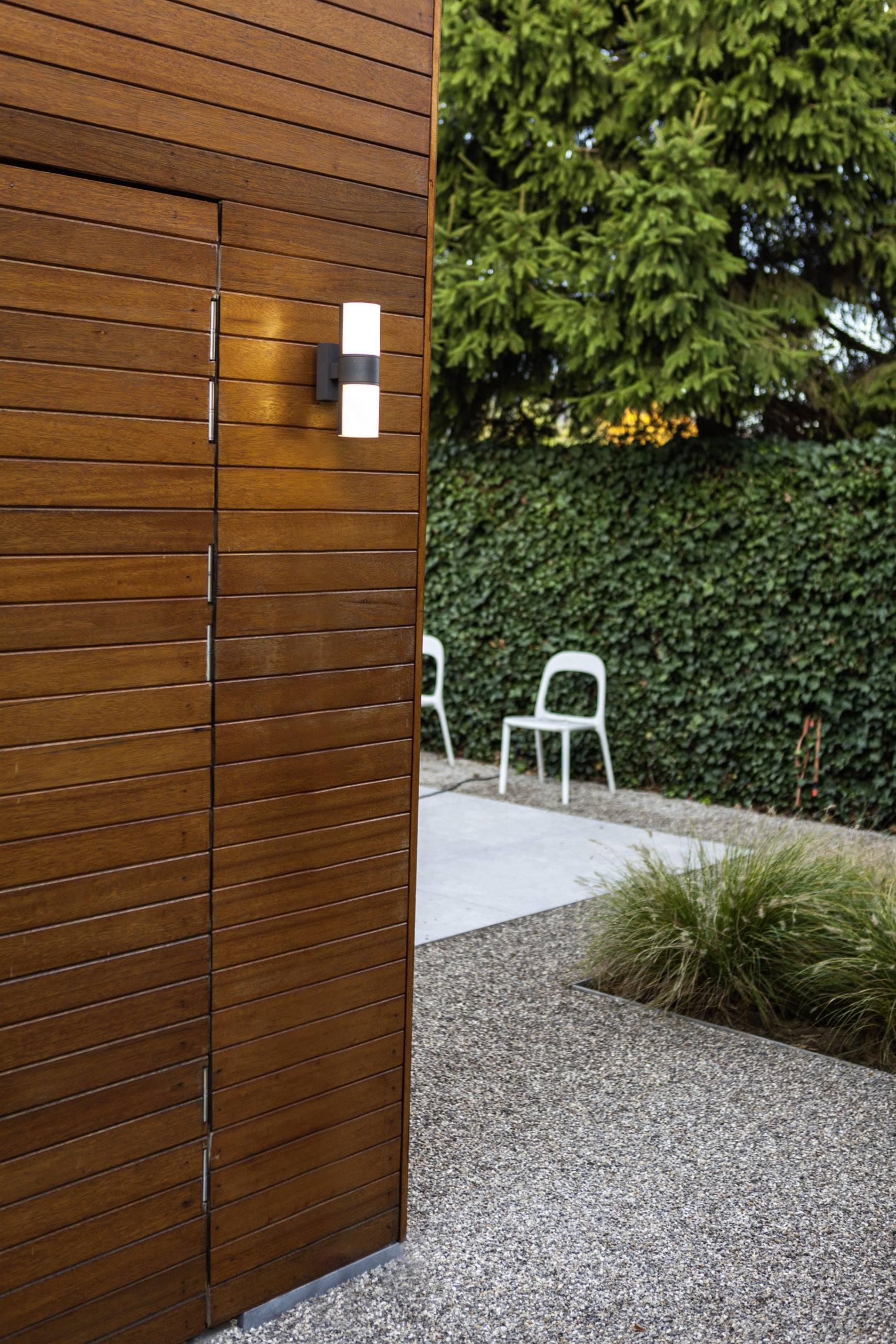 Mur en bois avec un éclairage extérieur, terrasse recouverte de gravier, deux chaises blanches à l'arrière, entourées de haies et de grands arbres.
