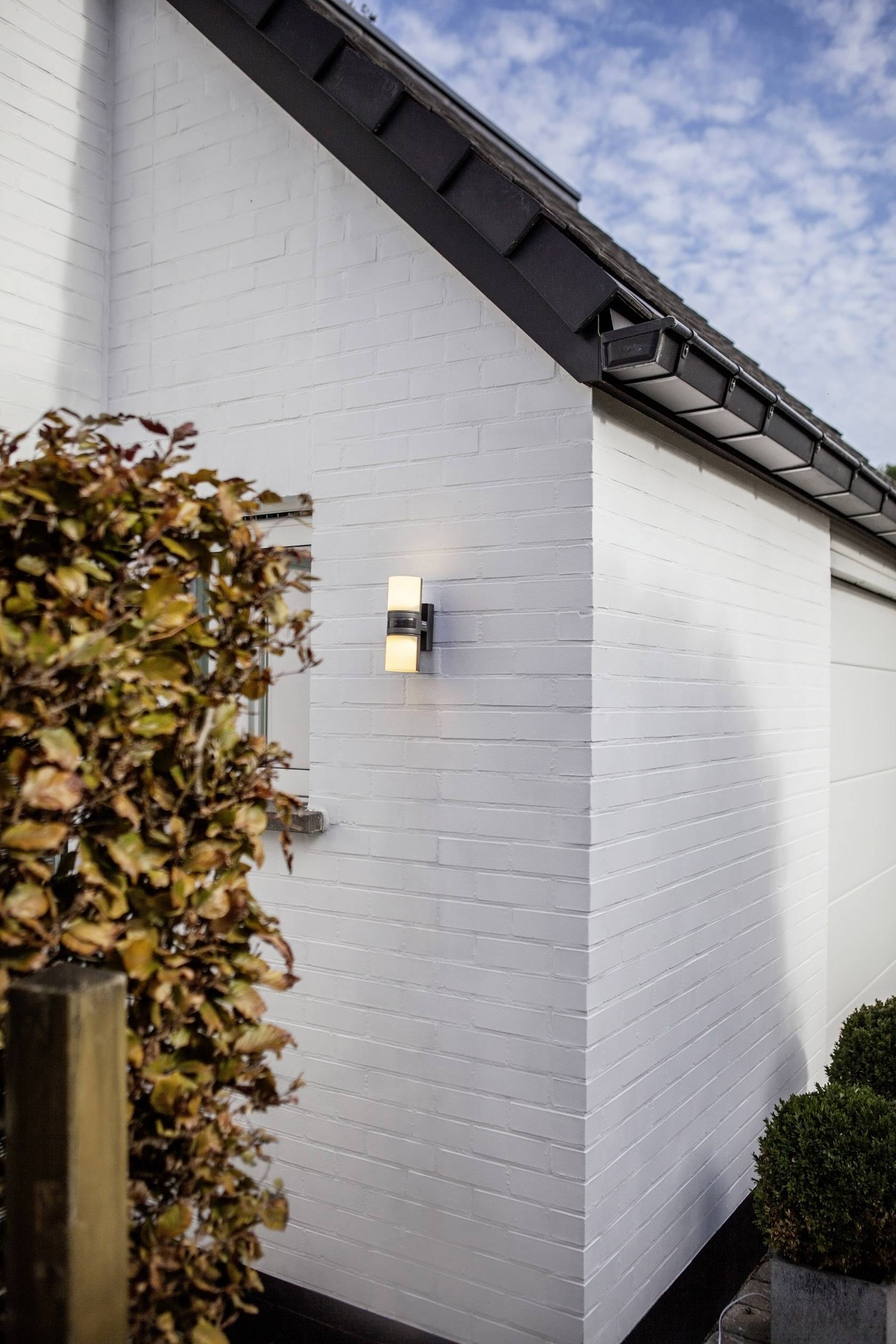 Mur de briques blanches d'une maison avec un luminaire extérieur moderne et lumineux ; à côté, des plantes automnales et un ciel bleu.