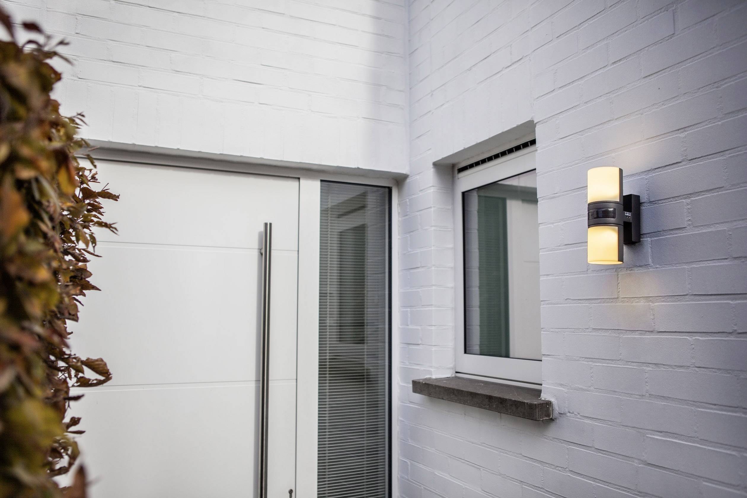 Façade blanche d'une maison avec une porte d'entrée moderne et un éclairage extérieur illuminé à côté d'une fenêtre.