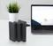 Sur une table, trois power banks noires sont posées devant un ordinateur portable ouvert affichant une image de paysage sur son écran ; à côté se trouve un pot de plante.