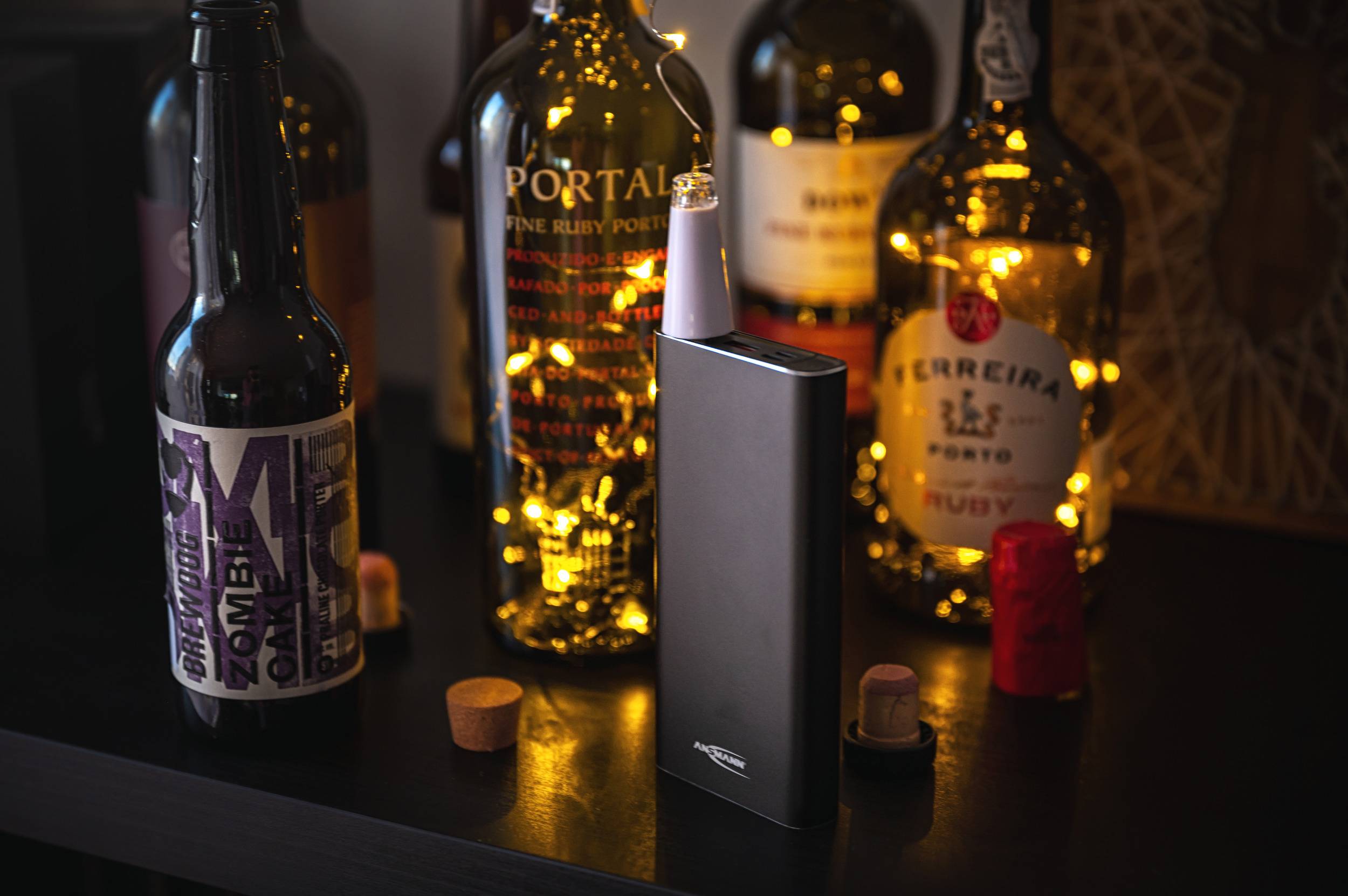 Bouteilles de vin et de bière sur une table, décorées avec des guirlandes lumineuses. Au premier plan, un appareil rectangulaire sombre.