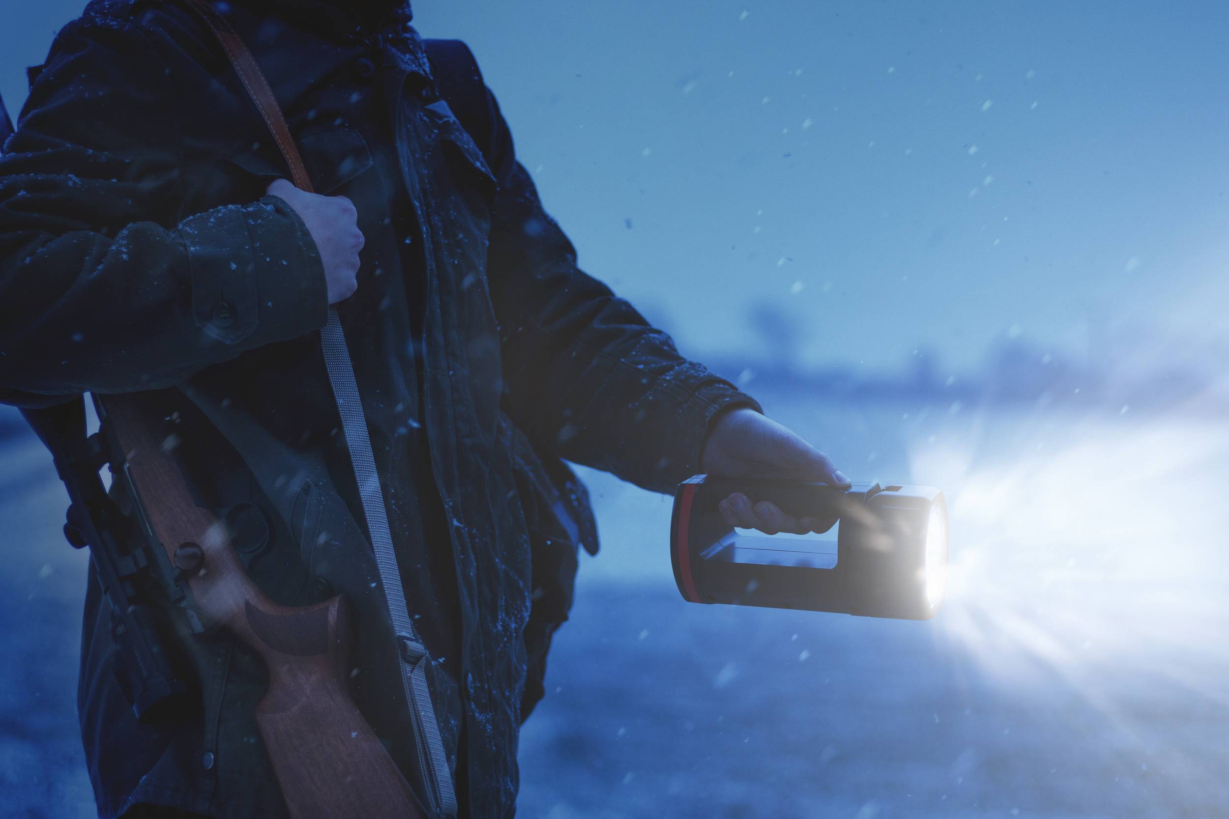 Personne avec un fusil de chasse et une lampe de poche dans la neige, illuminant les alentours à la tombée du jour.