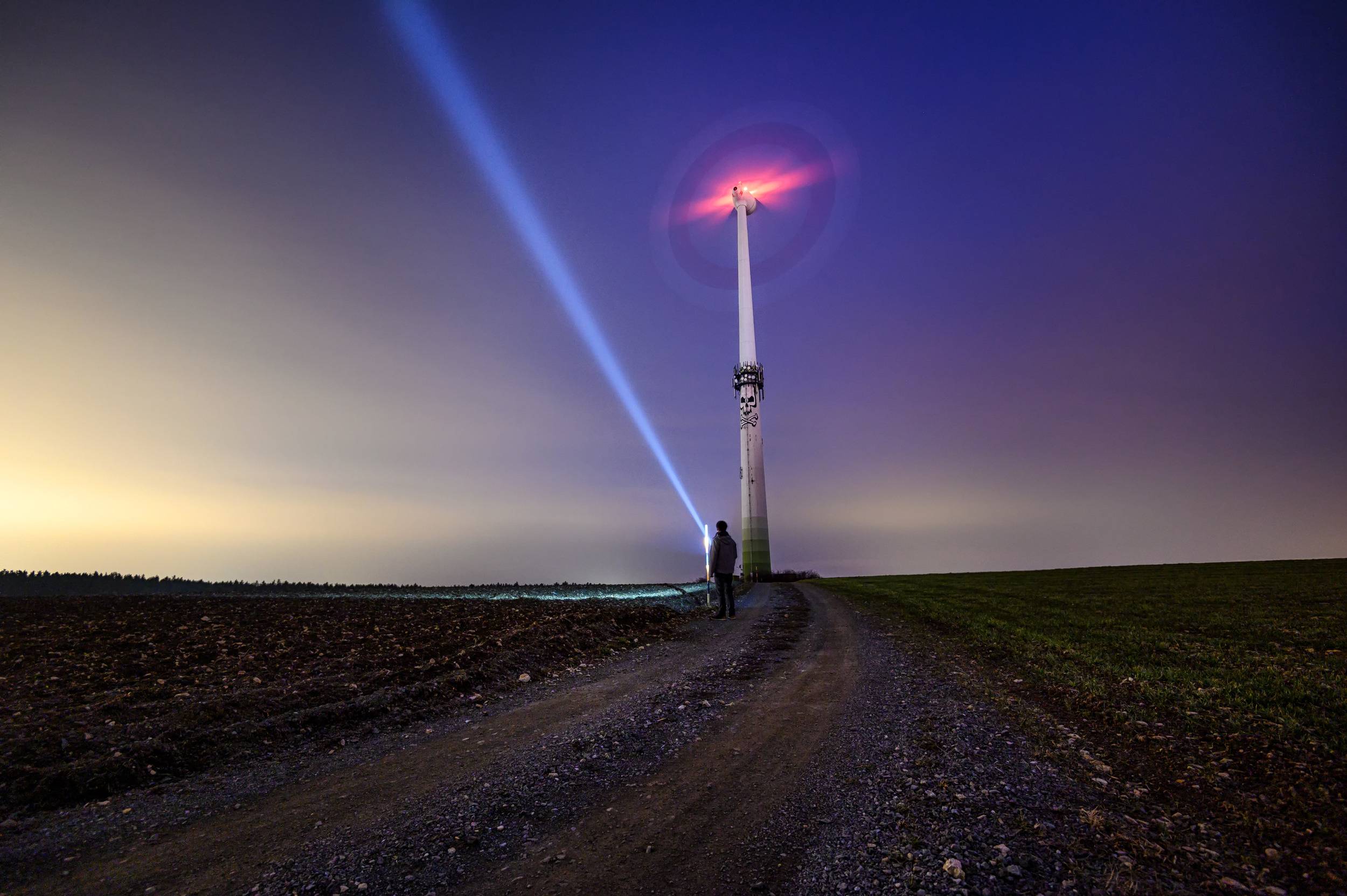 Une grande éolienne se dresse dans un paysage de fin de journée. Un faisceau lumineux bleu clair s'élève du sol en direction des rotors.