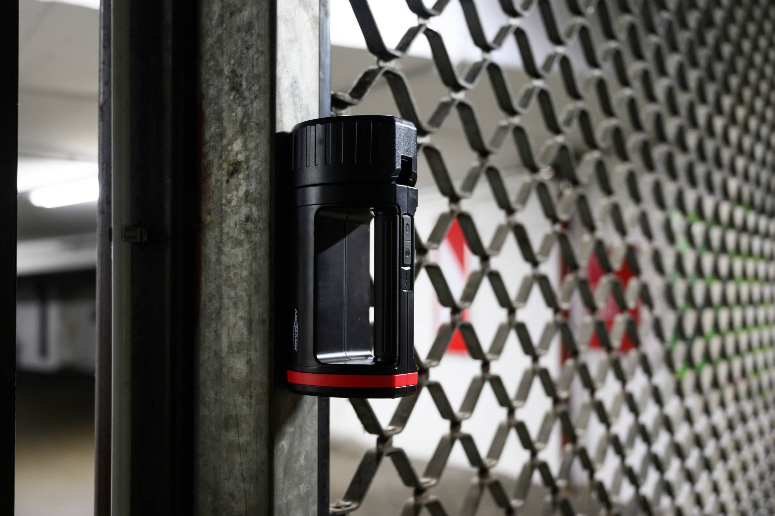 Une lampe de poche magnétique noire et rouge est fixée à une grille dans un parking. Des lumières floues sont visibles en arrière-plan.