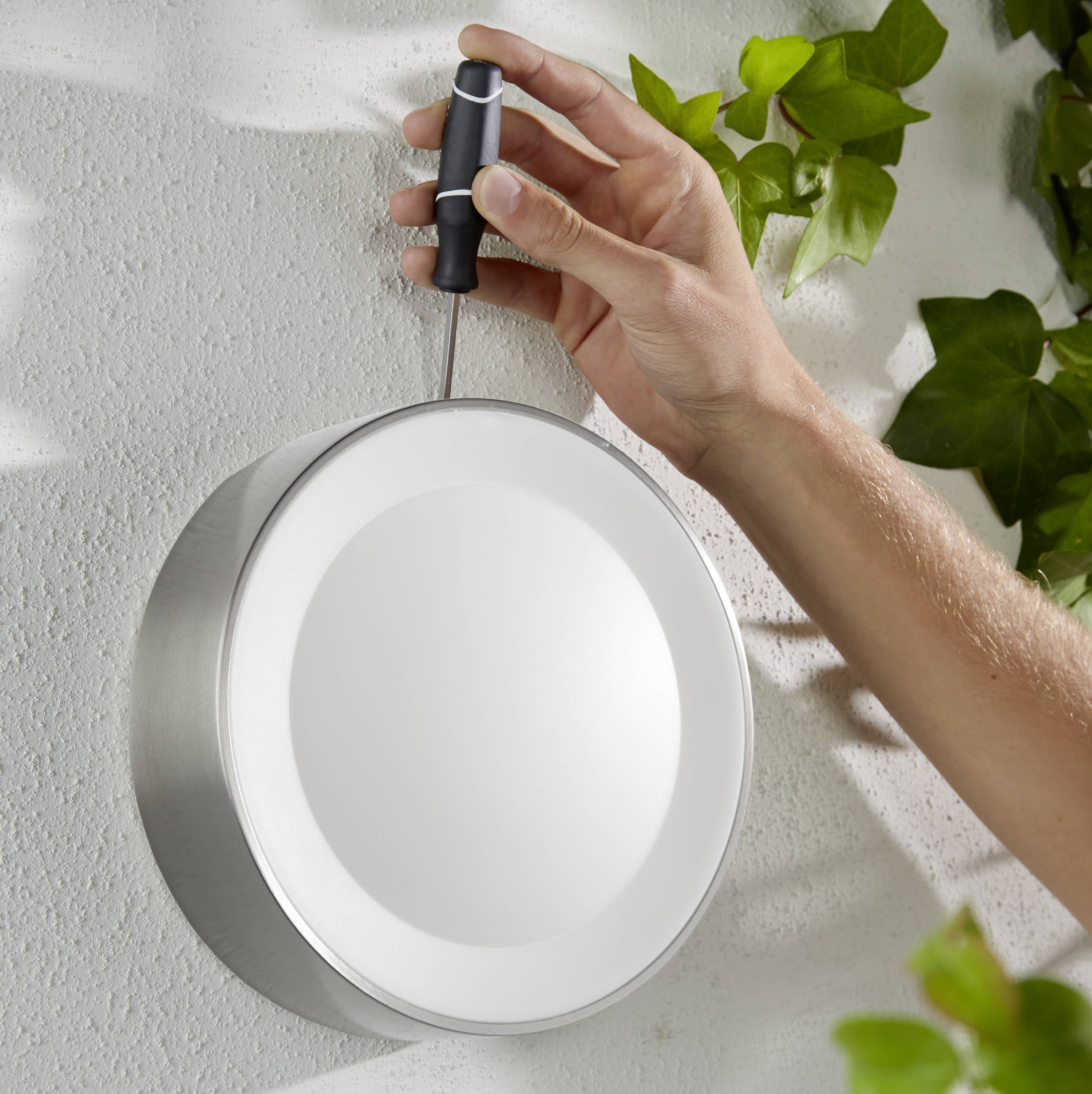 Une main tourne une vis sur un applique murale ronde en métal contre un mur orné de feuilles vertes.