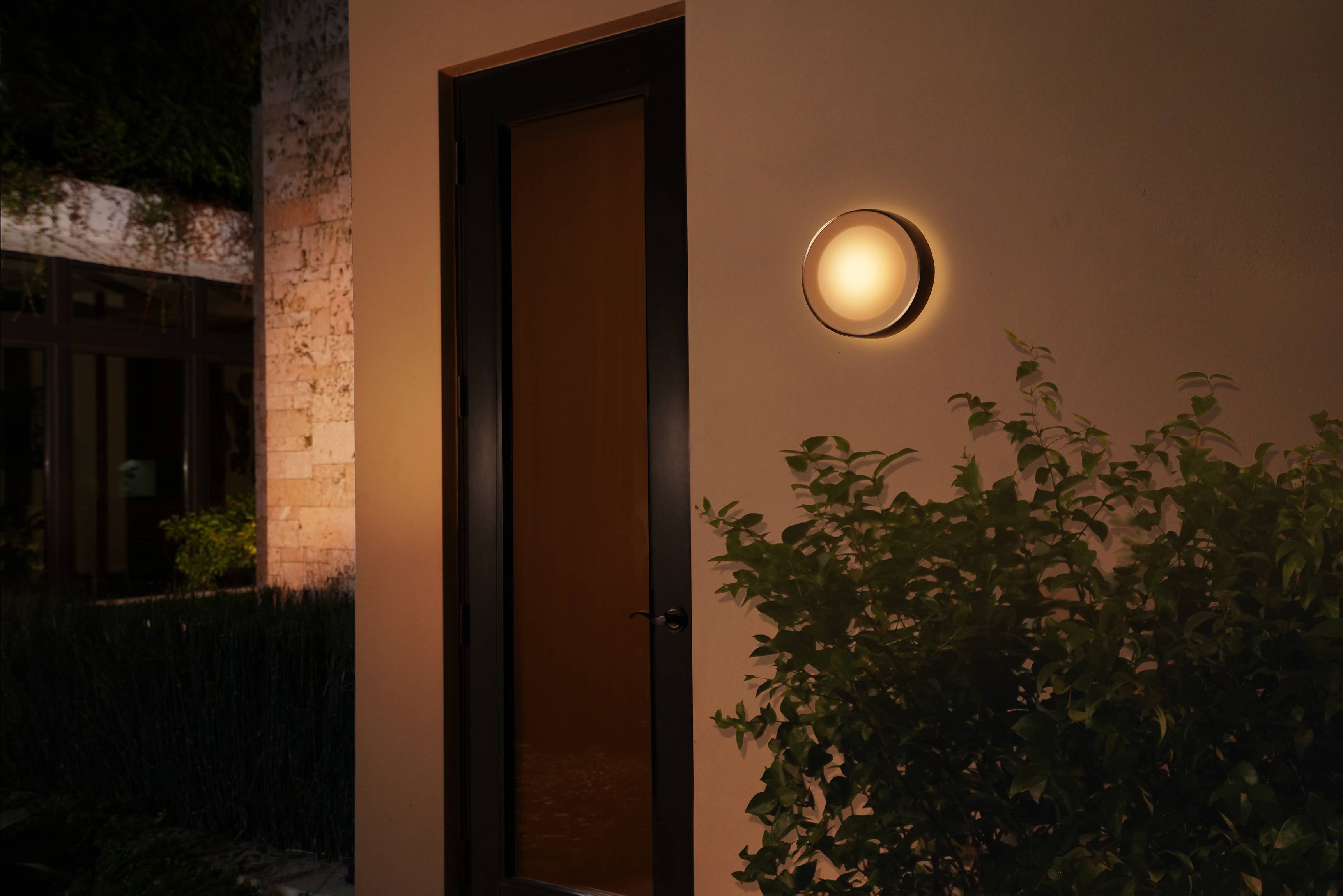 Un éclairage extérieur circulaire illuminé à côté d'une porte dans une scène de soirée, entouré de plantes et de lumière chaleureuse.