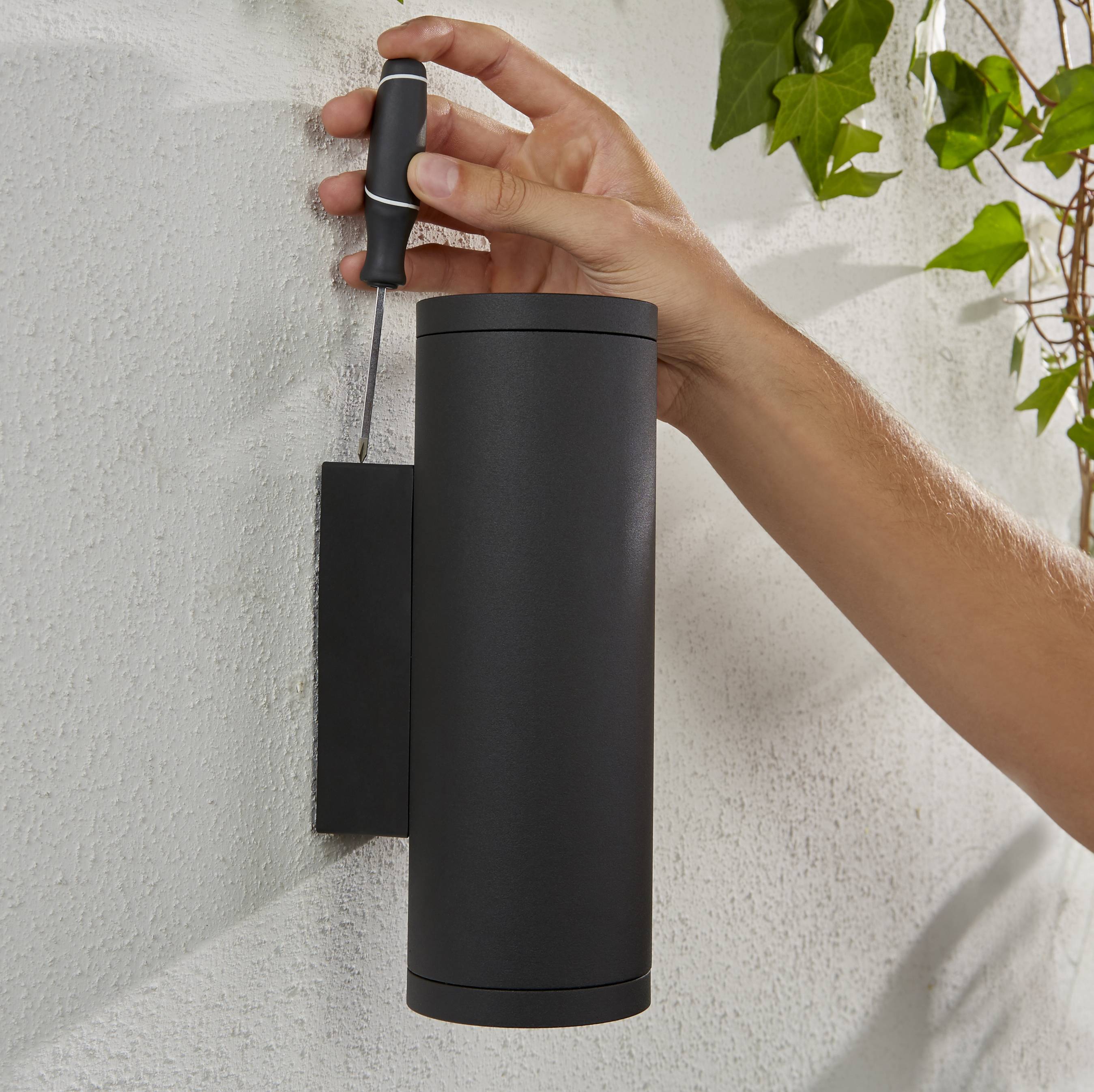 Une main tire sur une corde reliée à un cylindre noir fixé à un mur, entouré de feuilles vertes.