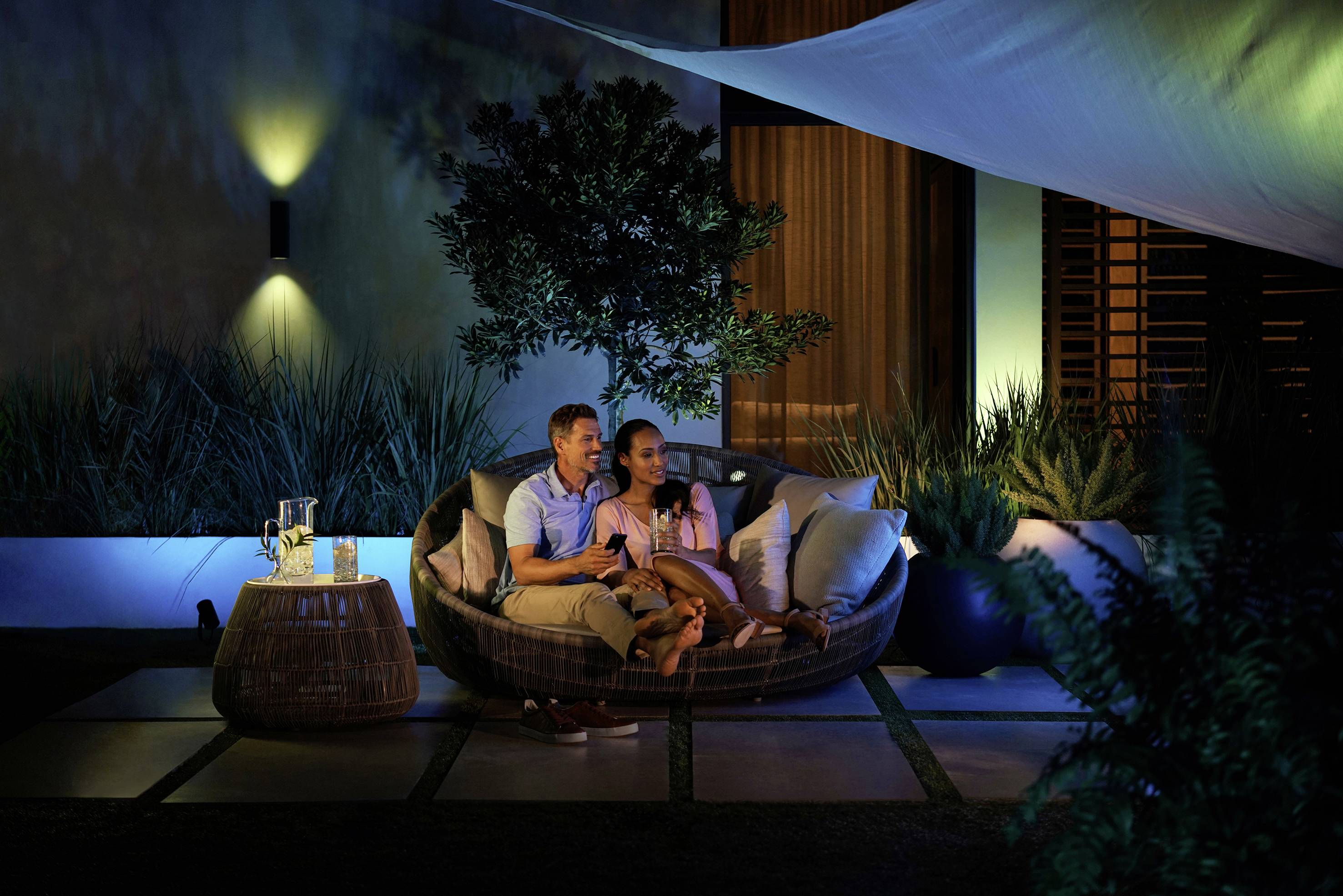Un couple est assis confortablement le soir sur une terrasse. Entourés de plantes et d'une lumière douce, ils se détendent sur un canapé rond.