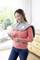 Une femme se tient dans une pièce, un coussin chauffant sur les épaules, tenant une télécommande et souriant. Une fenêtre est visible en arrière-plan.