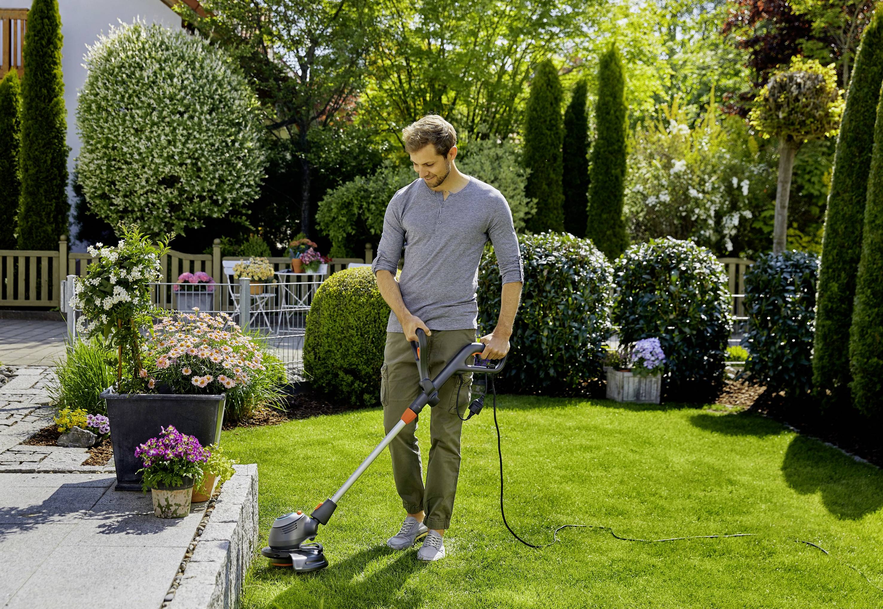 Un homme tond avec une tondeuse électrique la pelouse dans un jardin bien entretenu avec des fleurs et des arbustes par une journée ensoleillée.