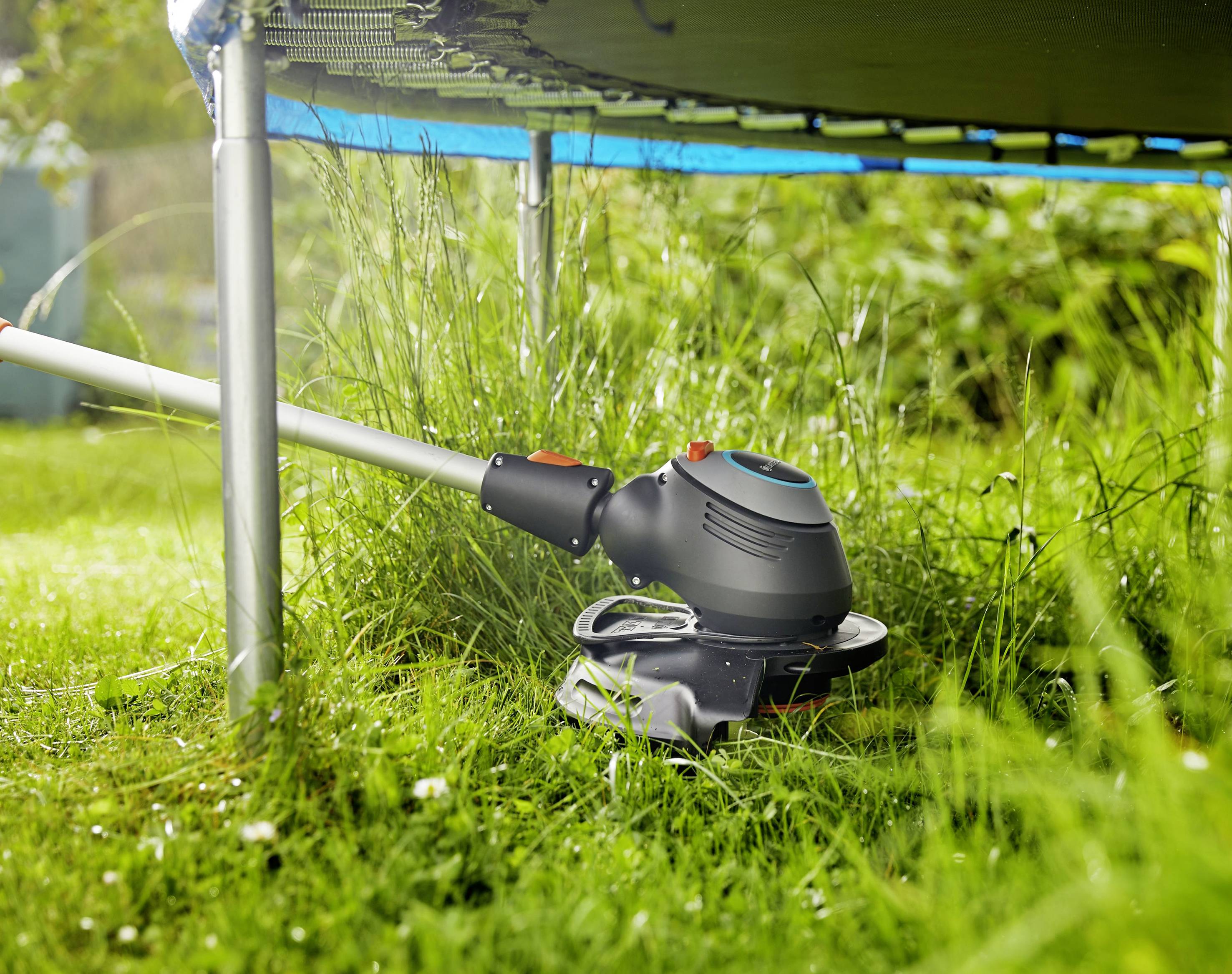 Une débroussailleuse électrique coupe l'herbe à côté d'un trampoline sur une pelouse verte. La lumière du soleil tombe sur la scène.