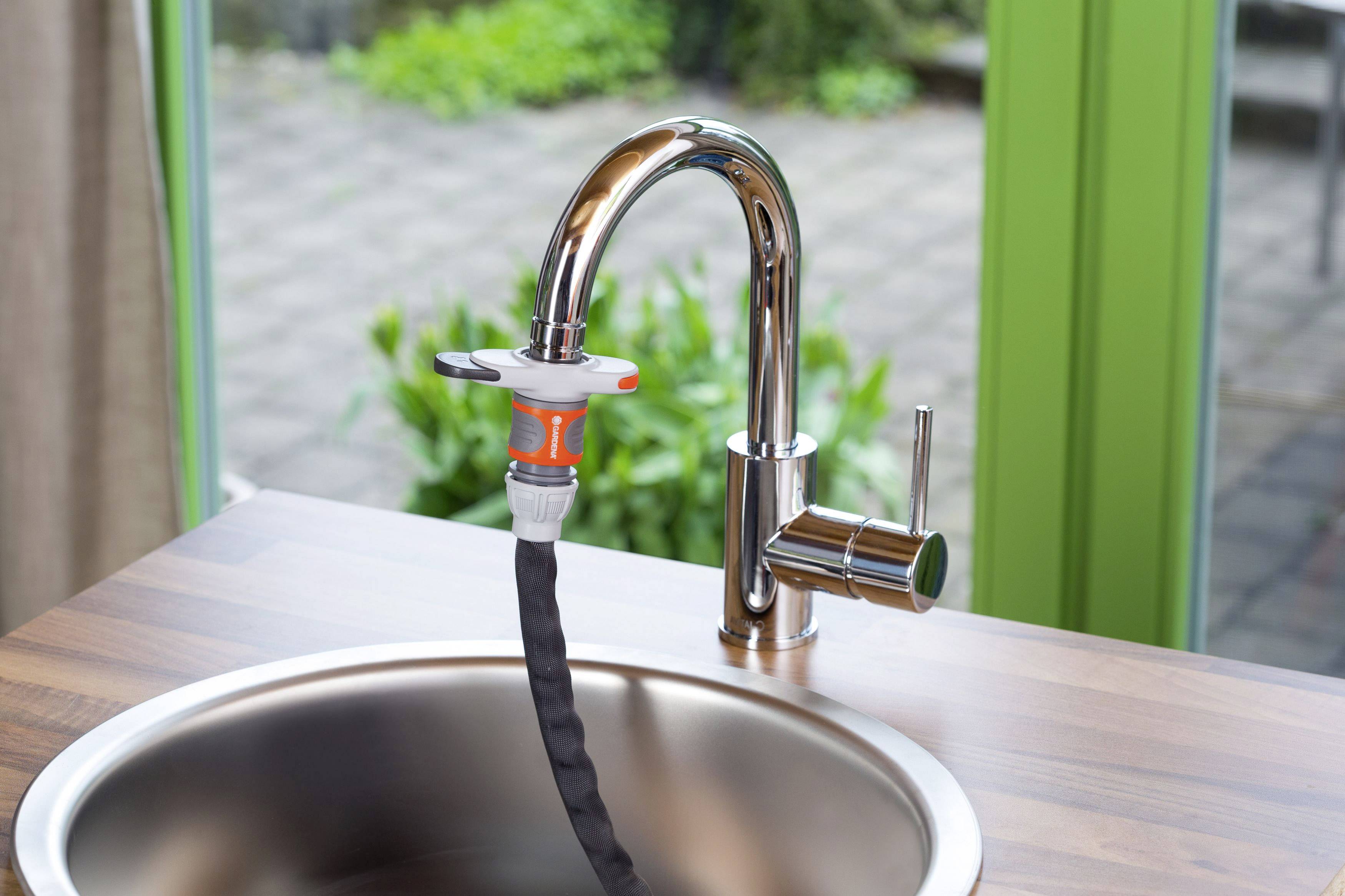 Un robinet de cuisine équipé d'un adaptateur de tuyau d'arrosage. À l'arrière-plan, on aperçoit des carreaux de jardin et des plantes vertes.