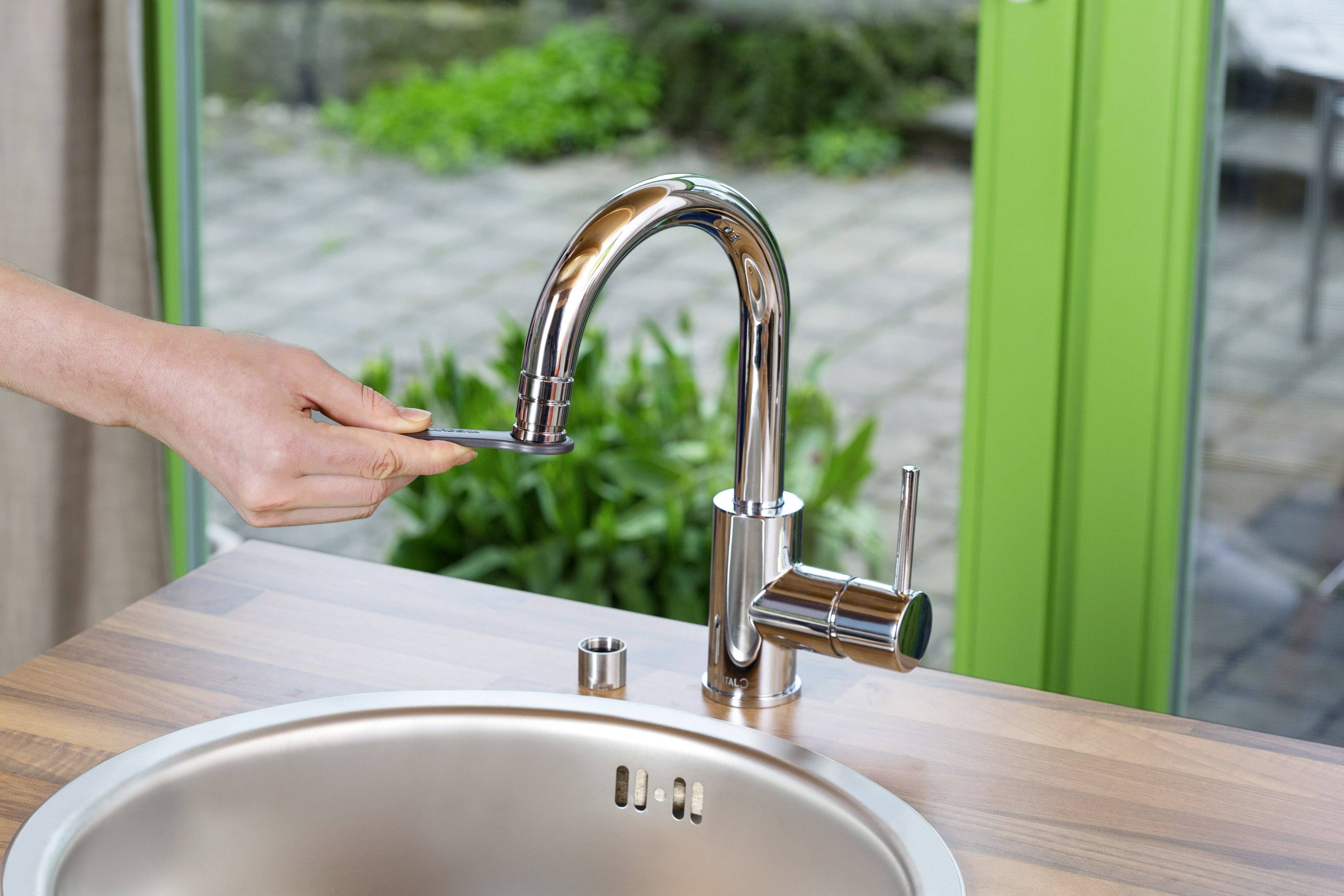 Une main actionne le levier d'un robinet de cuisine moderne sur un plan de travail en bois. En arrière-plan, une fenêtre donne sur un jardin.