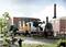 Une locomotive à vapeur miniature tire des wagons devant un bâtiment industriel. Des personnes et des arbres sont visibles en arrière-plan.