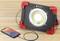 Lampe de travail LED portable avec cadre rouge, capable de charger un smartphone via un câble USB. Elle est posée sur une table en bois.