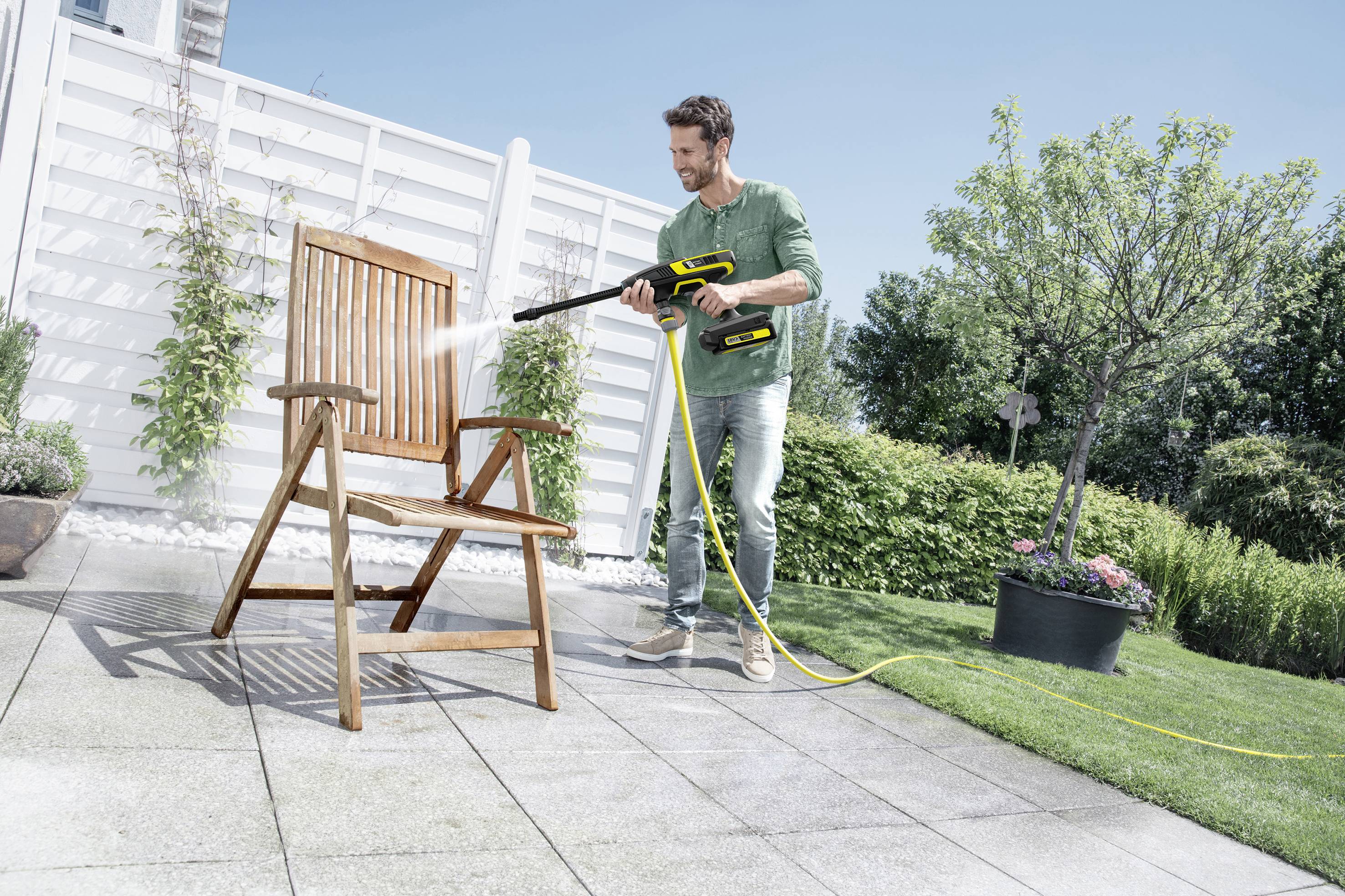 Un homme nettoie une chaise en bois avec un nettoyeur haute pression sur une terrasse de jardin par une journée ensoleillée.