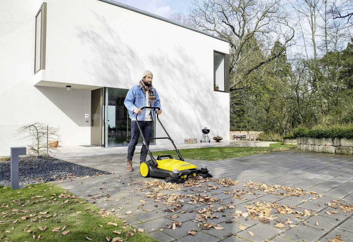Une personne utilise un balai-poussoir jaune pour dégager les feuilles de l'allée d'une maison moderne, entourée d'arbres et de verdure.