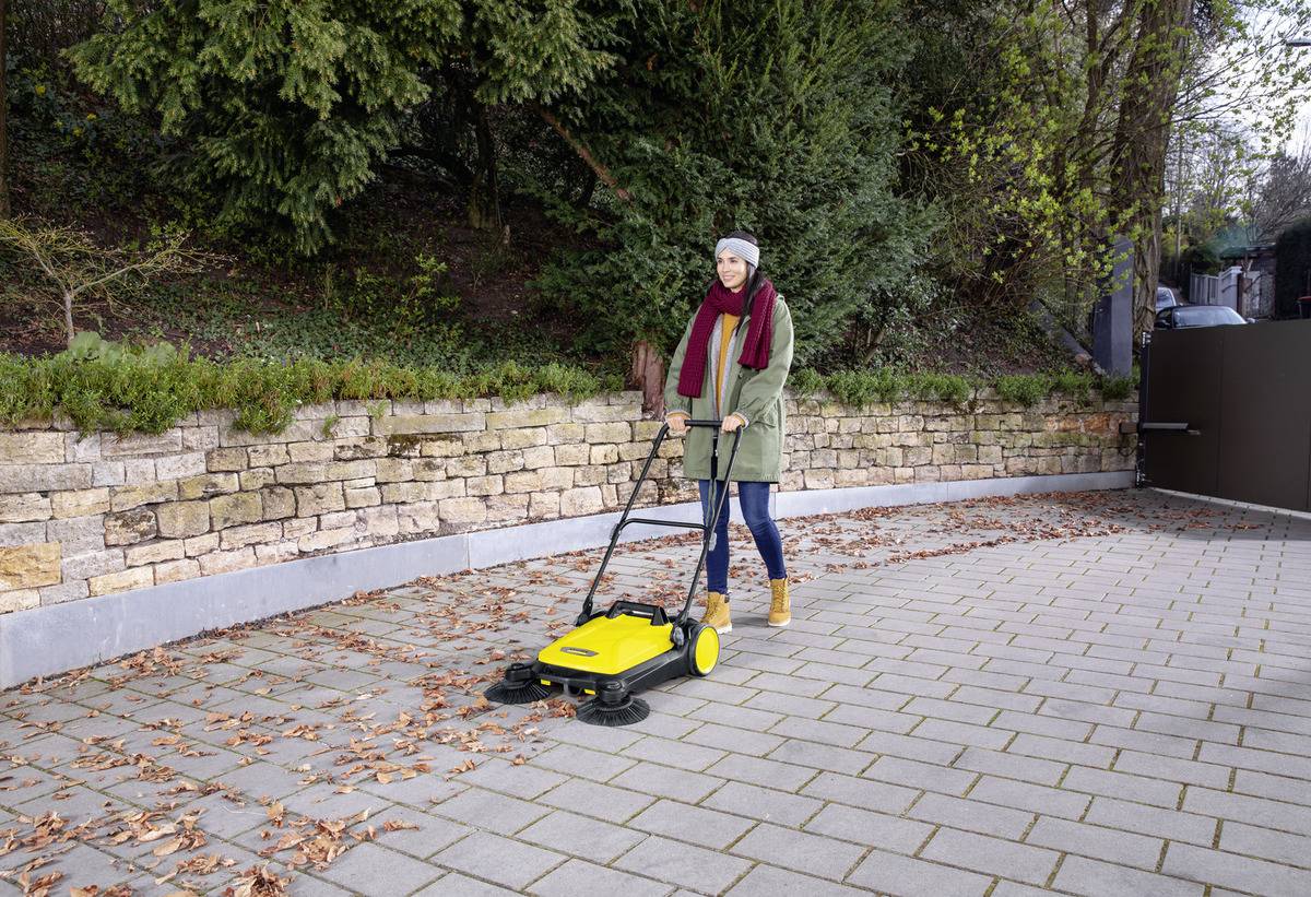 Une personne balayant des feuilles sur un chemin pavé à l'aide d'un balai-poussoir jaune. La scène se déroule à l'extérieur, avec de la verdure et un mur en pierre en arrière-plan.