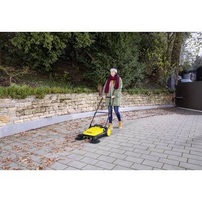 Une personne balayant des feuilles sur un chemin pavé à l'aide d'un balai-poussoir jaune. La scène se déroule à l'extérieur, avec de la verdure et un mur en pierre en arrière-plan.